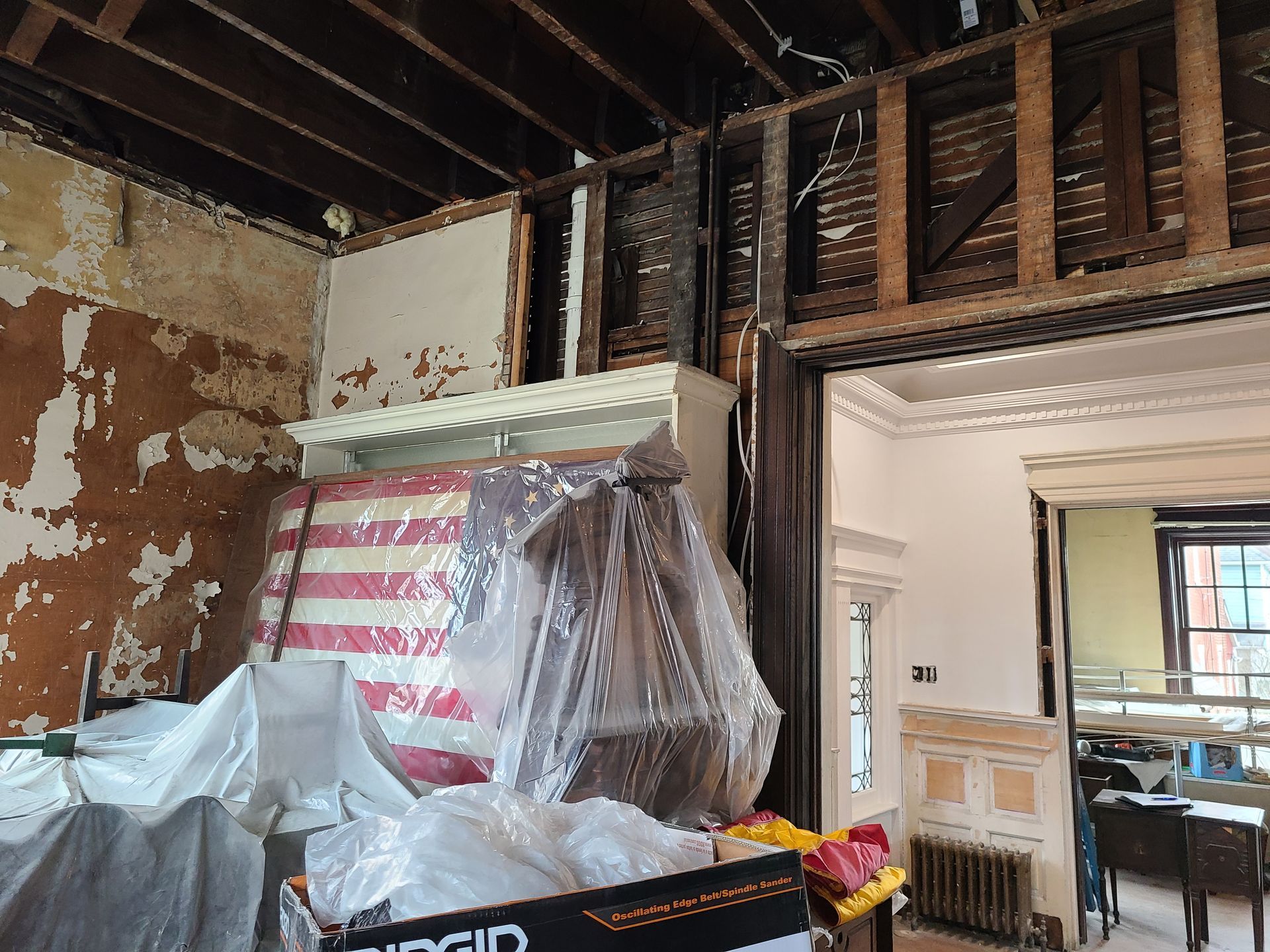 Room in disrepair, walls stripped, exposed wood beams. Debris, furniture covered in plastic. Ornate doorway visible.
