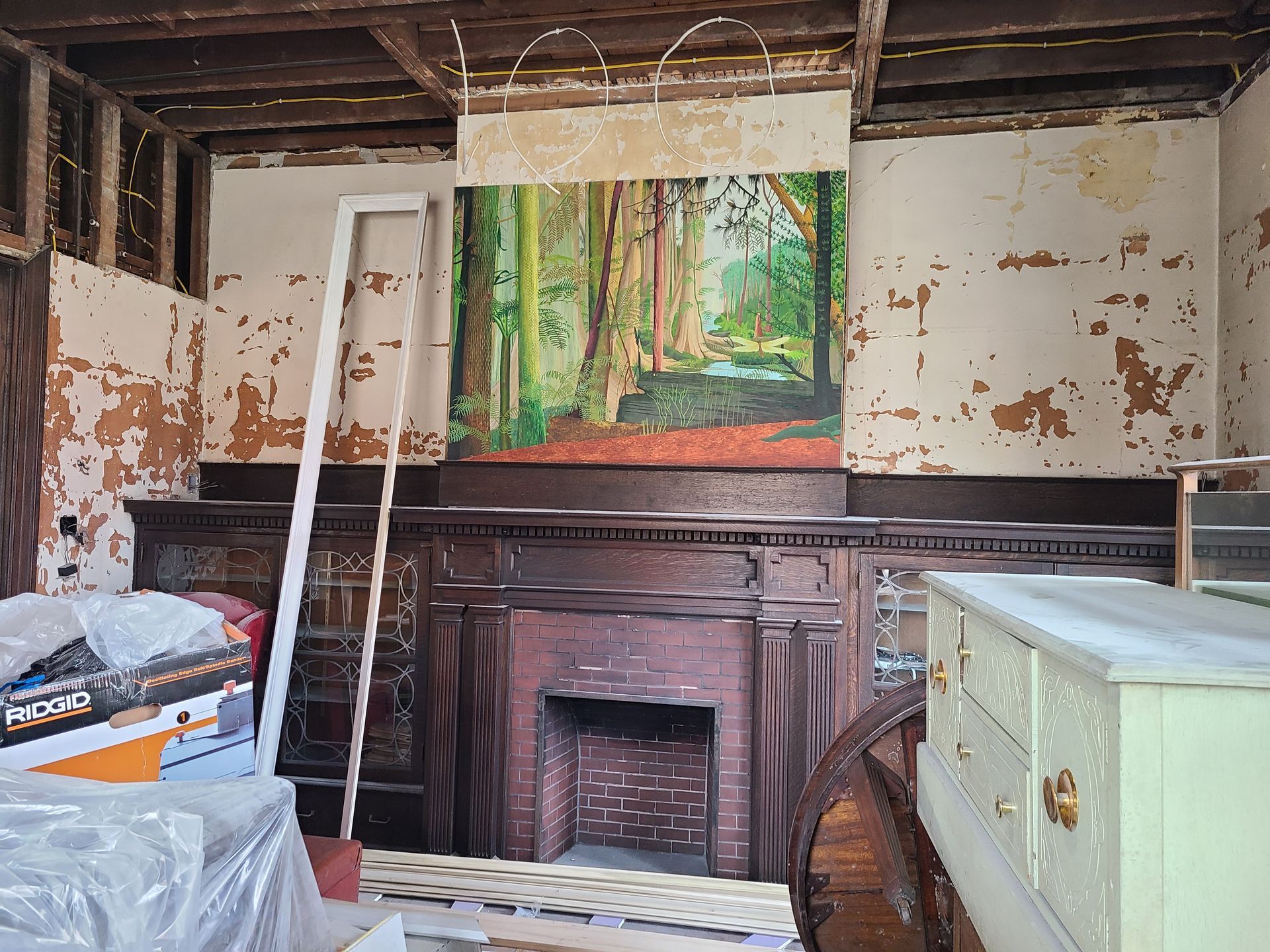 Room under renovation, showing a fireplace with a painting of a forest and peeling paint on the walls.