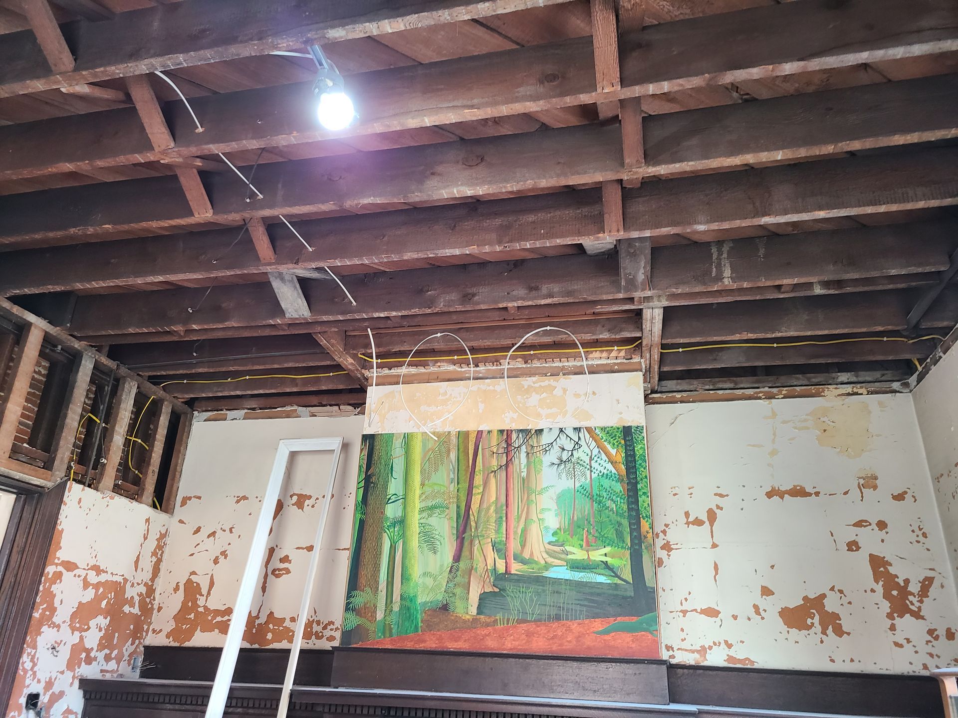 Room under renovation; exposed ceiling beams, peeling paint, large forest painting on the wall, and ladder.