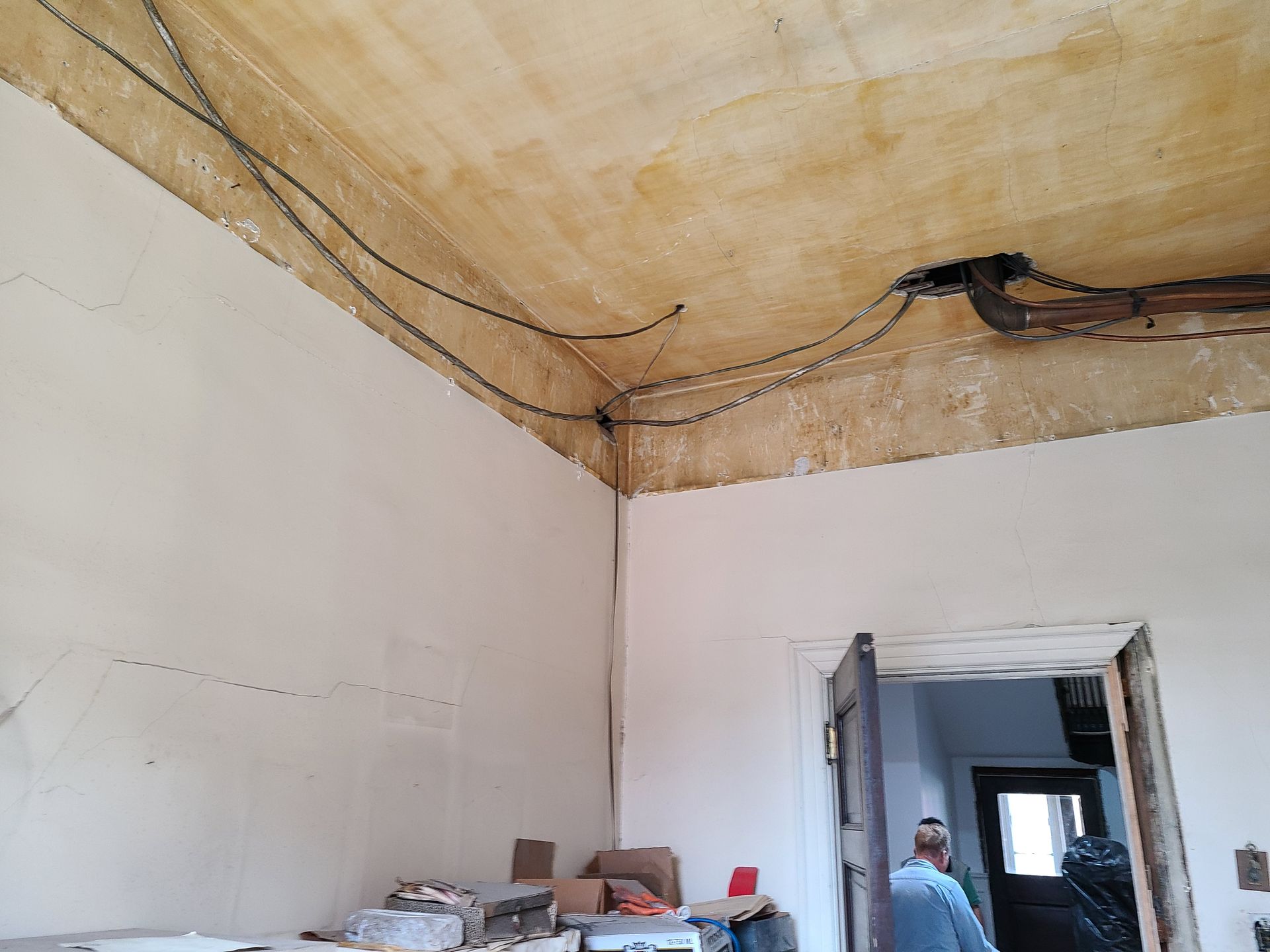 Room with exposed wiring on ceiling, walls partially plastered. A person stands in a doorway.