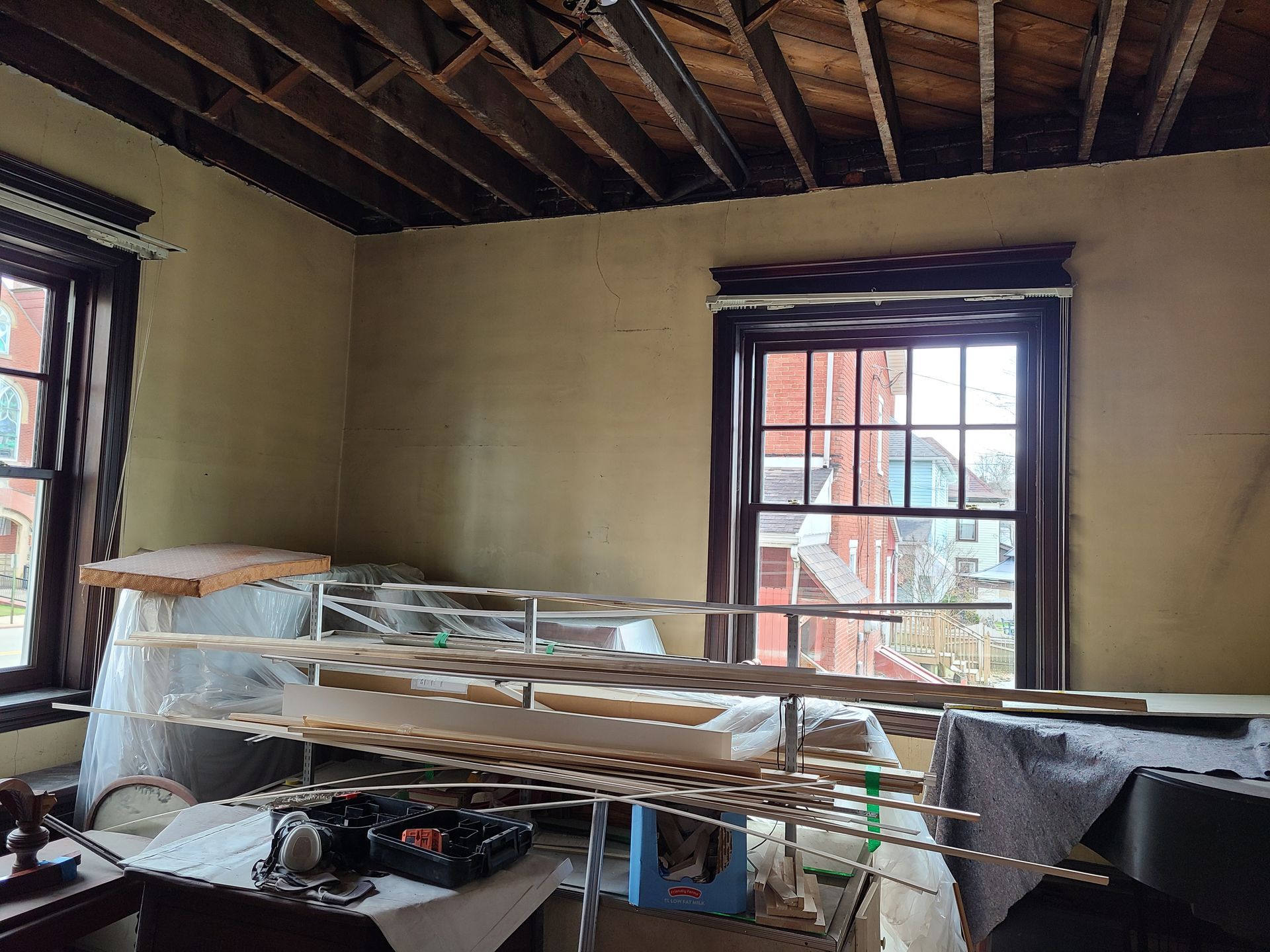 Room with exposed ceiling beams and two windows, cluttered with shelves and items.