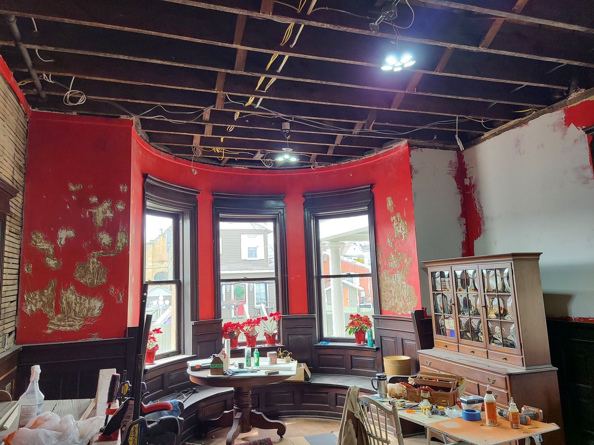 Interior room under renovation, red walls, bay window, exposed ceiling beams, furniture, and debris.