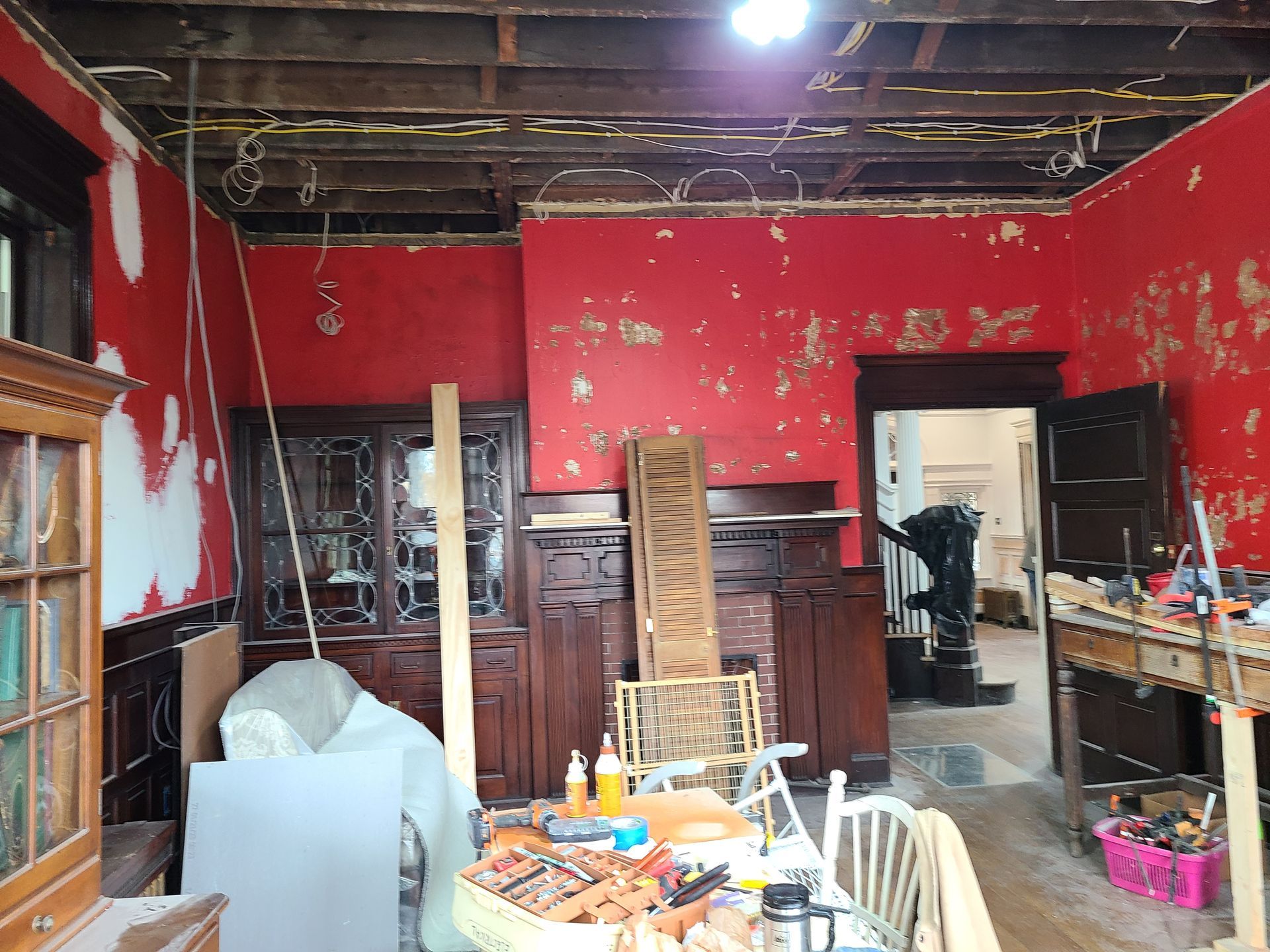 Room interior undergoing renovation, red walls with peeling paint, exposed ceiling beams, furniture, and tools scattered.