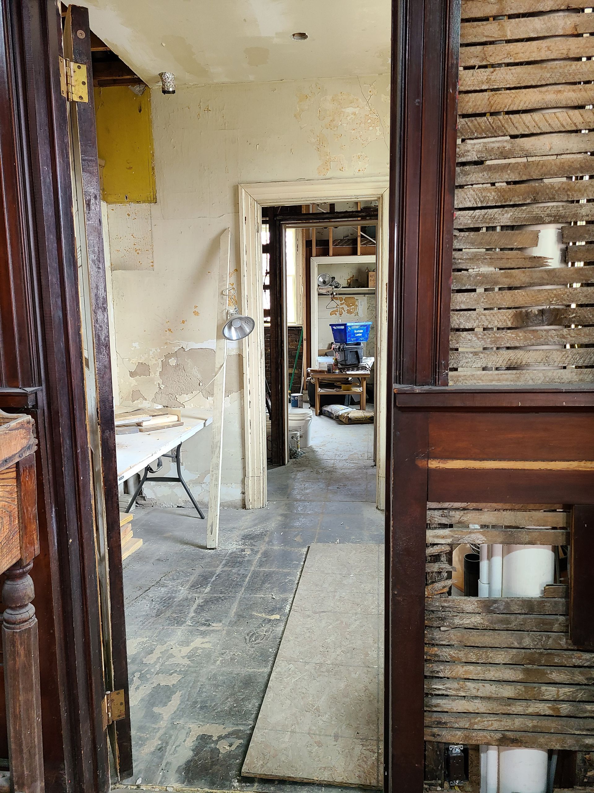 Interior hallway in disrepair. Exposed wood, plaster, and debris. Visible doorway leads to a cluttered room.