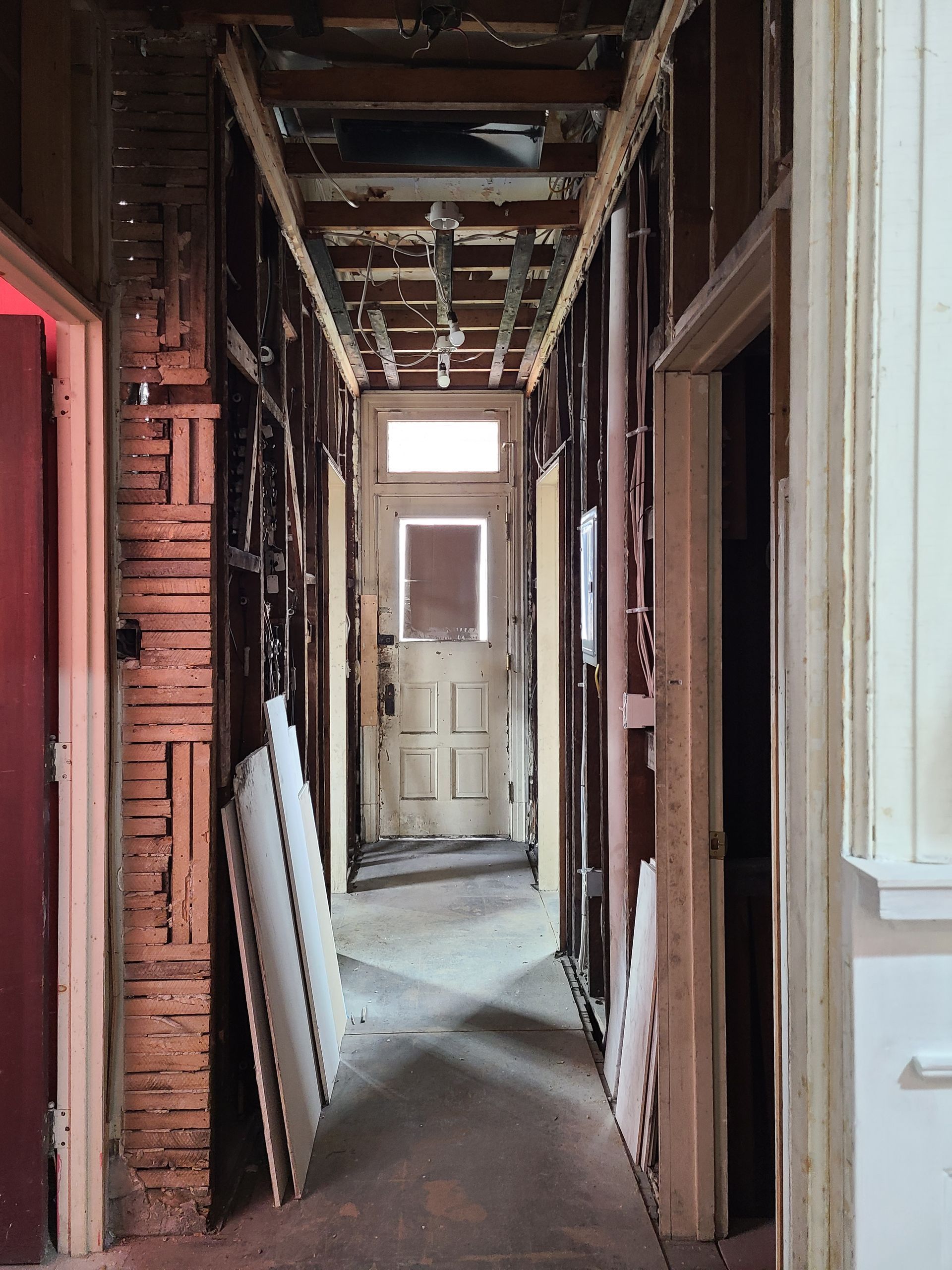 Hallway under renovation; exposed studs, partial drywall, doorway at the end with daylight.