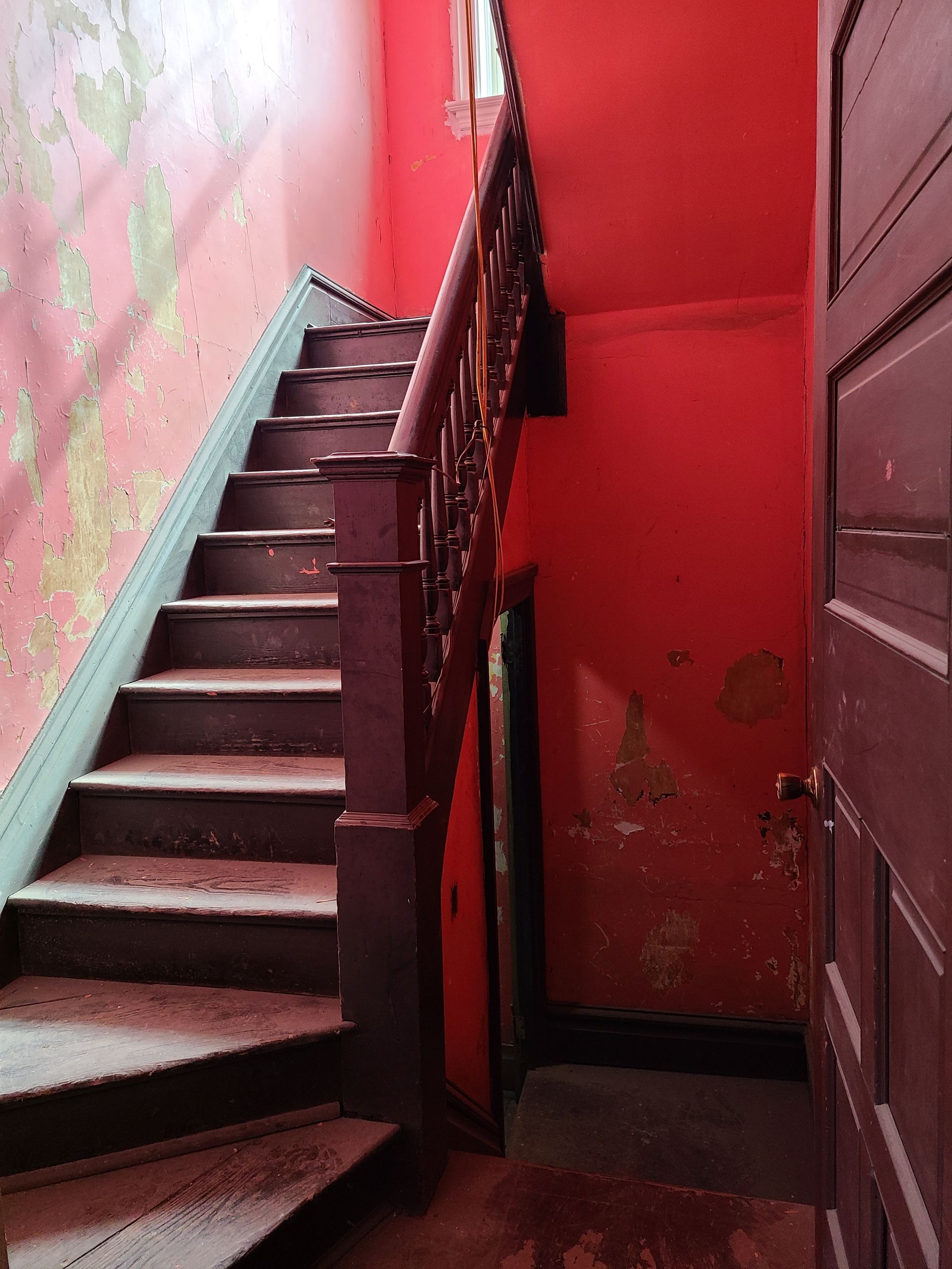 Wooden staircase in a red-painted hallway with peeling paint. A dark door is on the right.