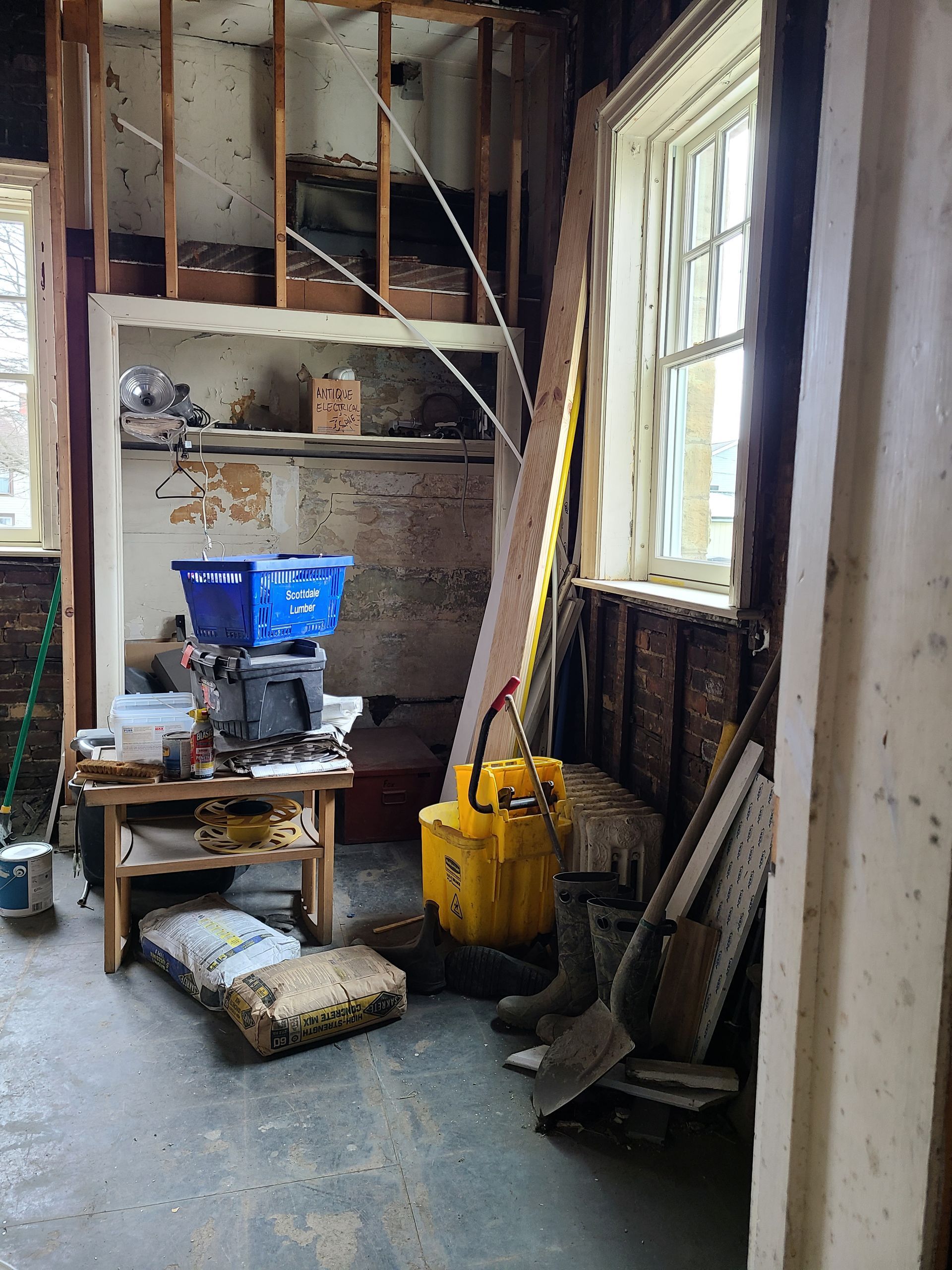 Interior renovation: walls exposed, work table with equipment, yellow power tool, two windows.