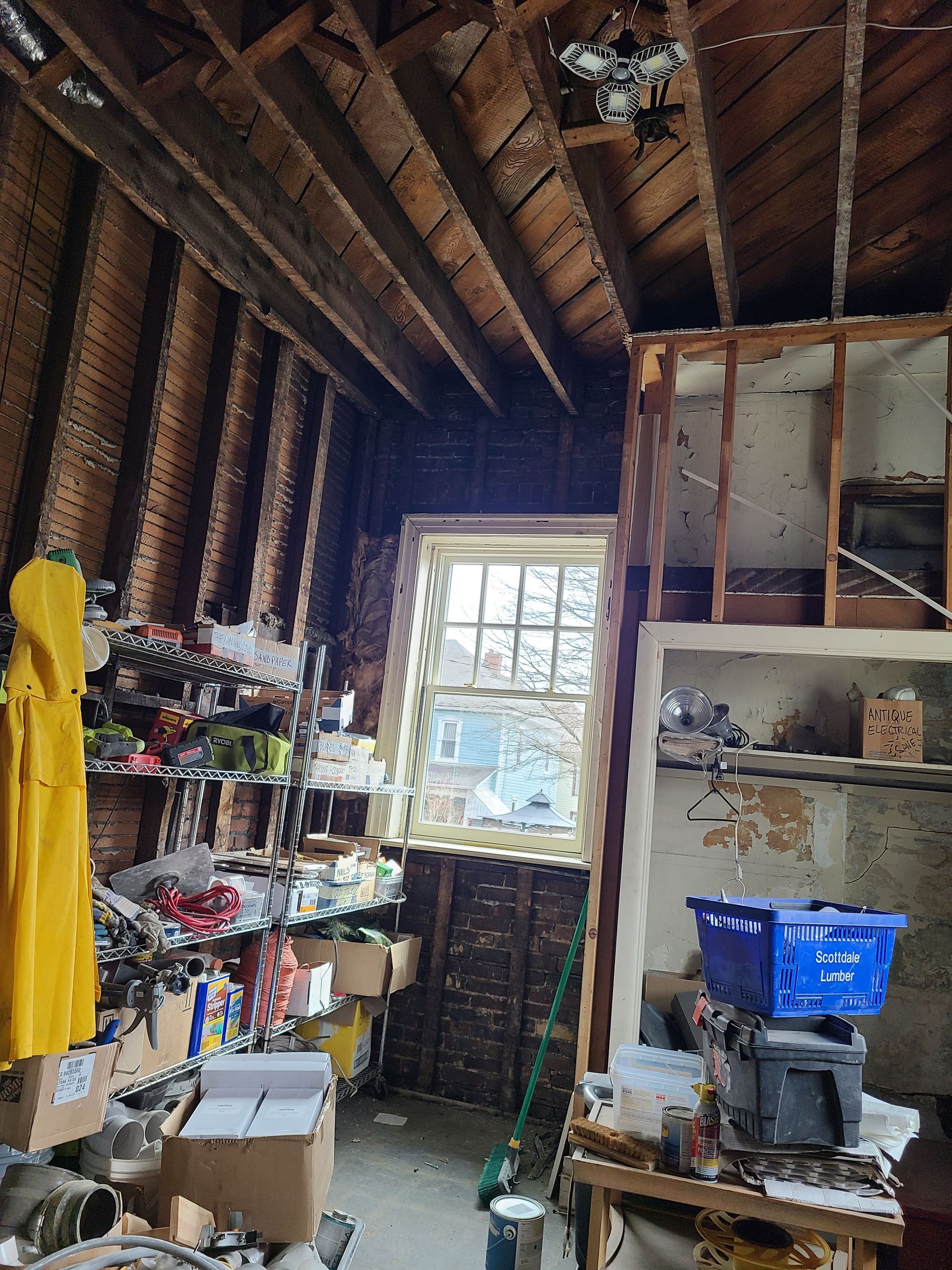 Cluttered attic space with exposed beams, window, and haphazard shelving filled with items.