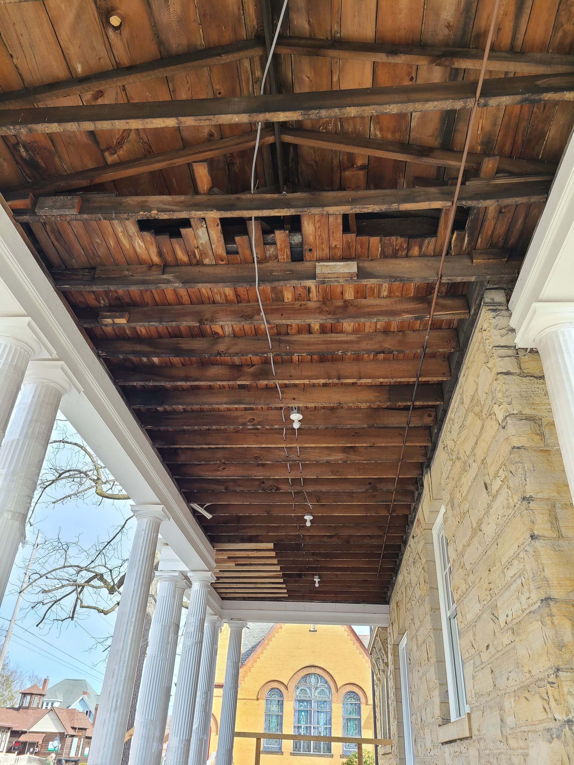 Damaged wooden porch ceiling with missing sections, viewed from below; building with columns and stone facade in background.