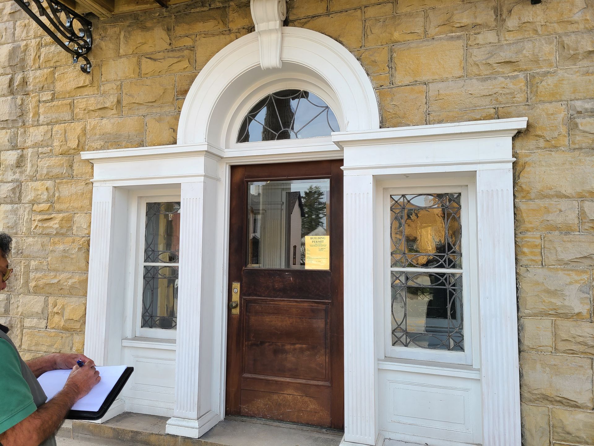 Person with clipboard examining a brown wooden door with a rounded top and two adjacent windows in a yellow stone building.