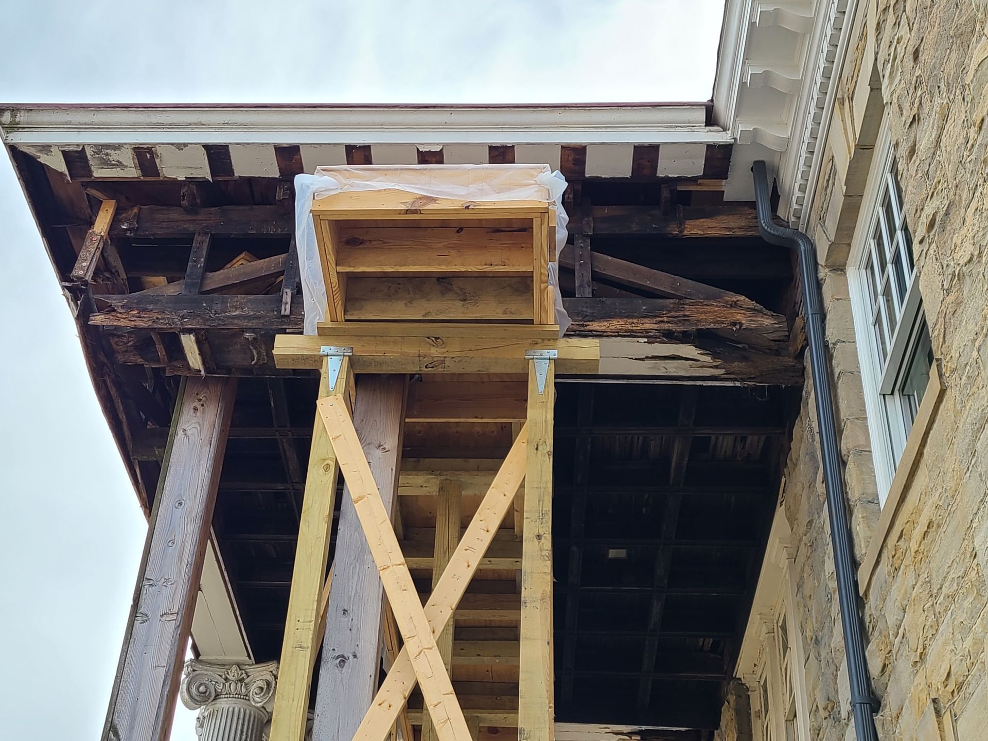 Exterior building undergoing repair. Wooden scaffolding supports a damaged section of a roof with exposed beams.