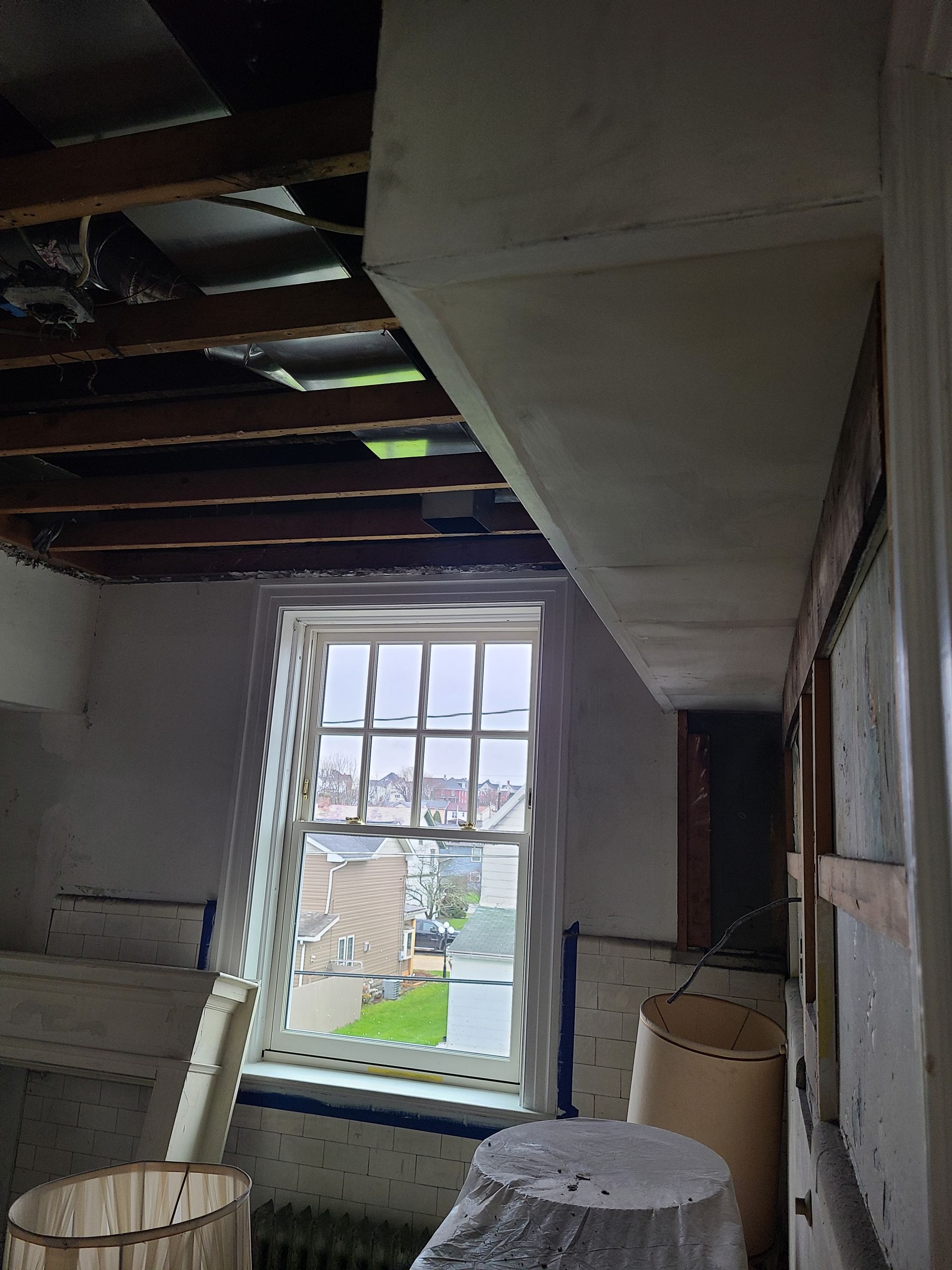 Interior view of a room under construction with exposed ceiling beams, a window, and construction debris.