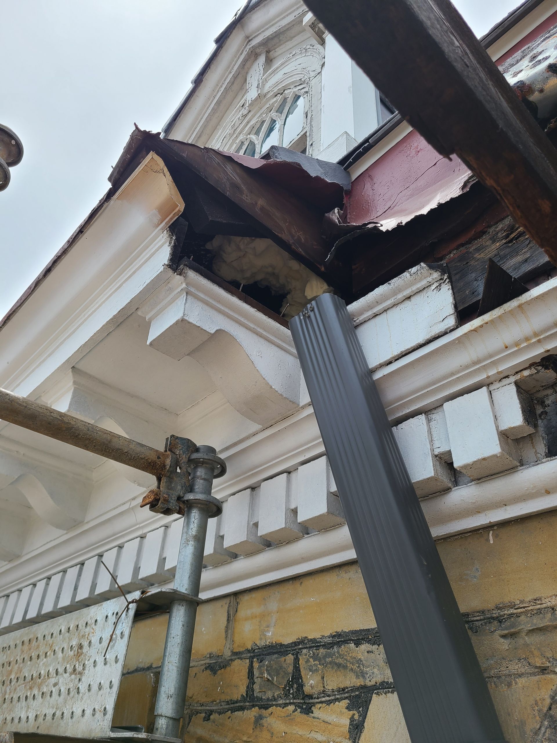 Damaged house corner with scaffolding and guttering; white trim, brick wall, and dark brown damage.