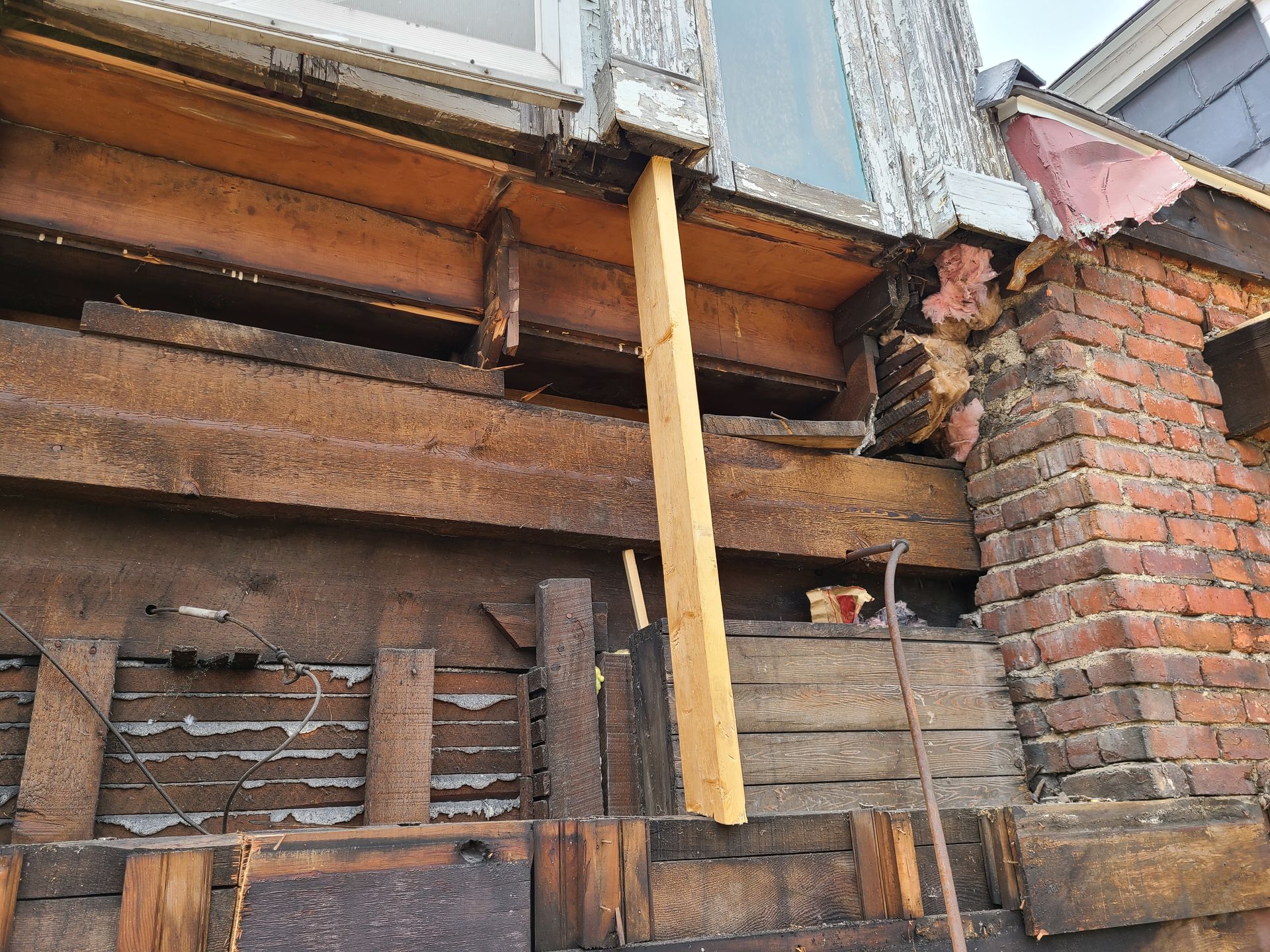 Exterior house wall damage with exposed beams, brick, and window. A wooden support prop is in place.