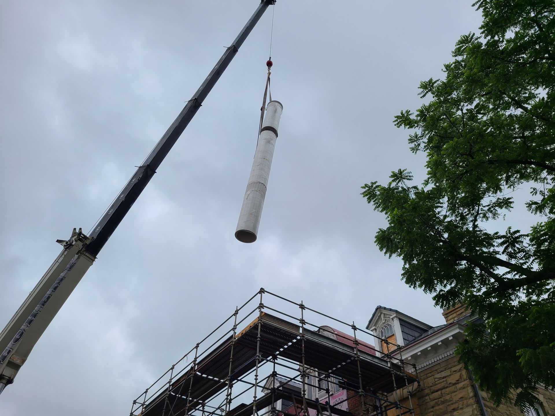 Crane lifting a cylindrical object near a building with scaffolding under a cloudy sky.