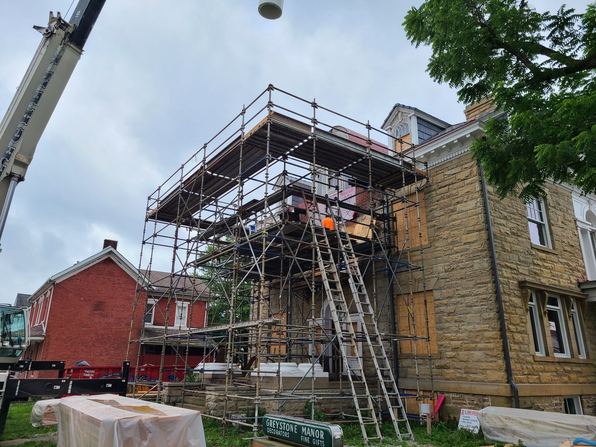 Scaffolding surrounds a stone house under construction, with a crane in the background. Cloudy sky.