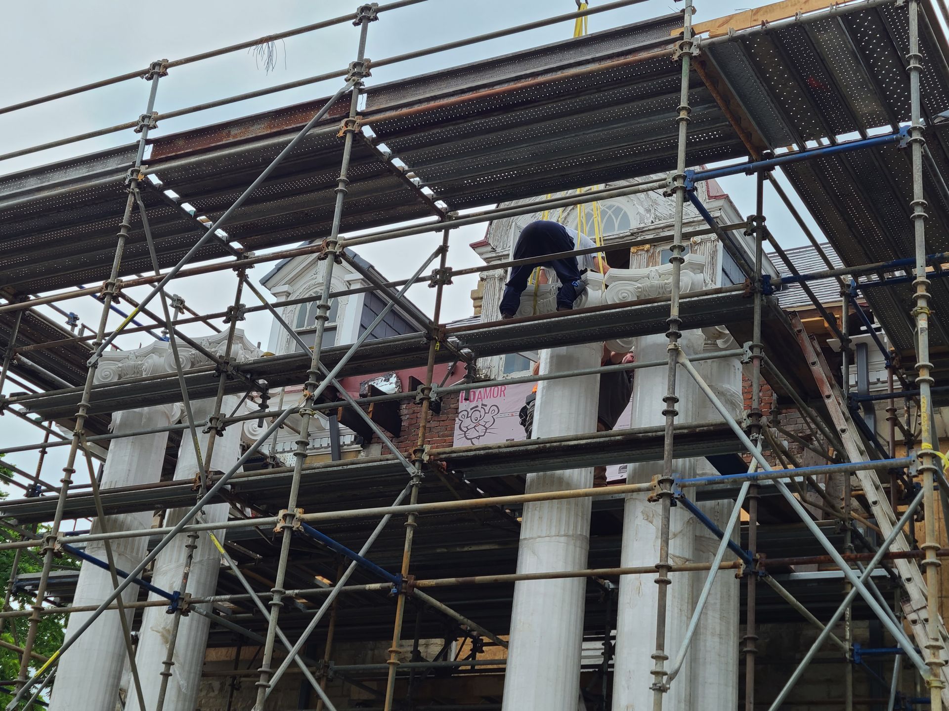 Building facade under construction, scaffolding surrounds. A person works on the roof. White columns visible.