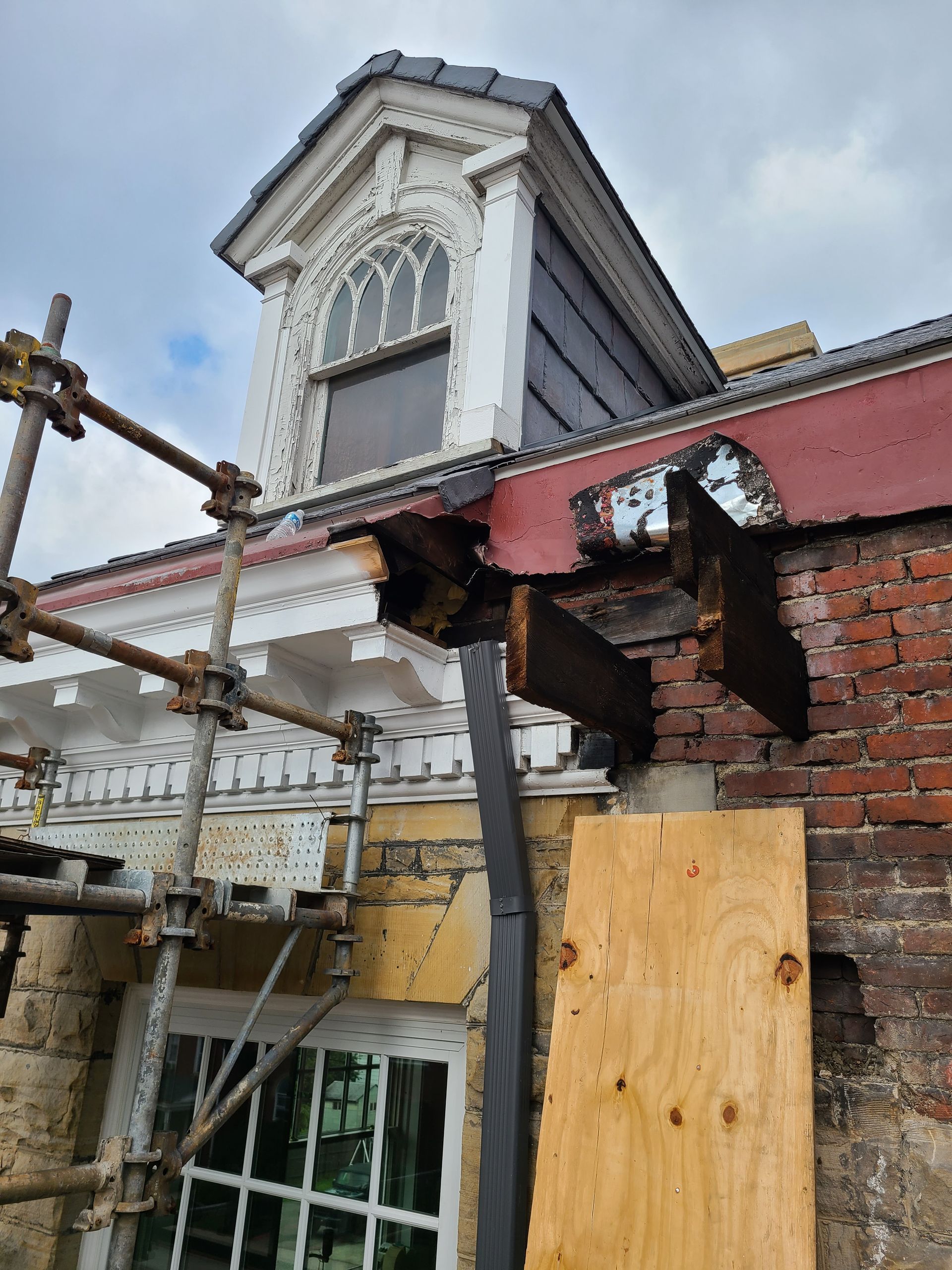 Damaged building exterior with exposed wood, red brick, scaffolding, and a dormer window.