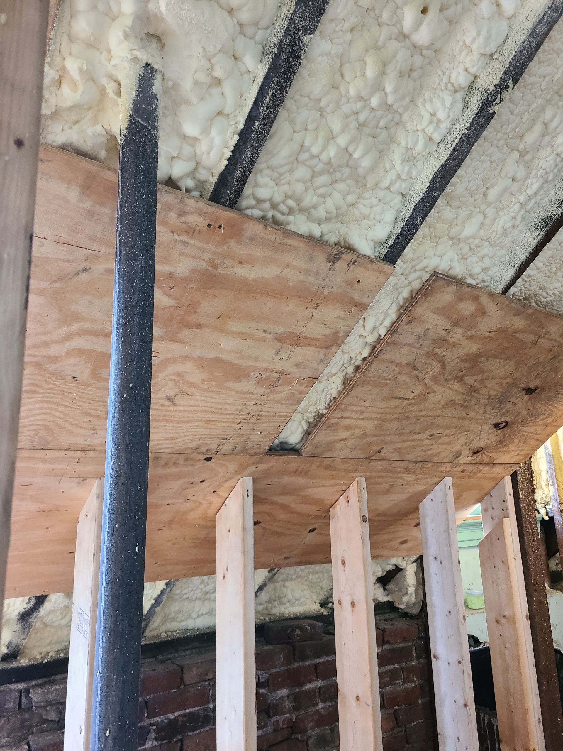 Construction detail: Plywood and beams with spray foam insulation visible. A black pipe extends upwards.