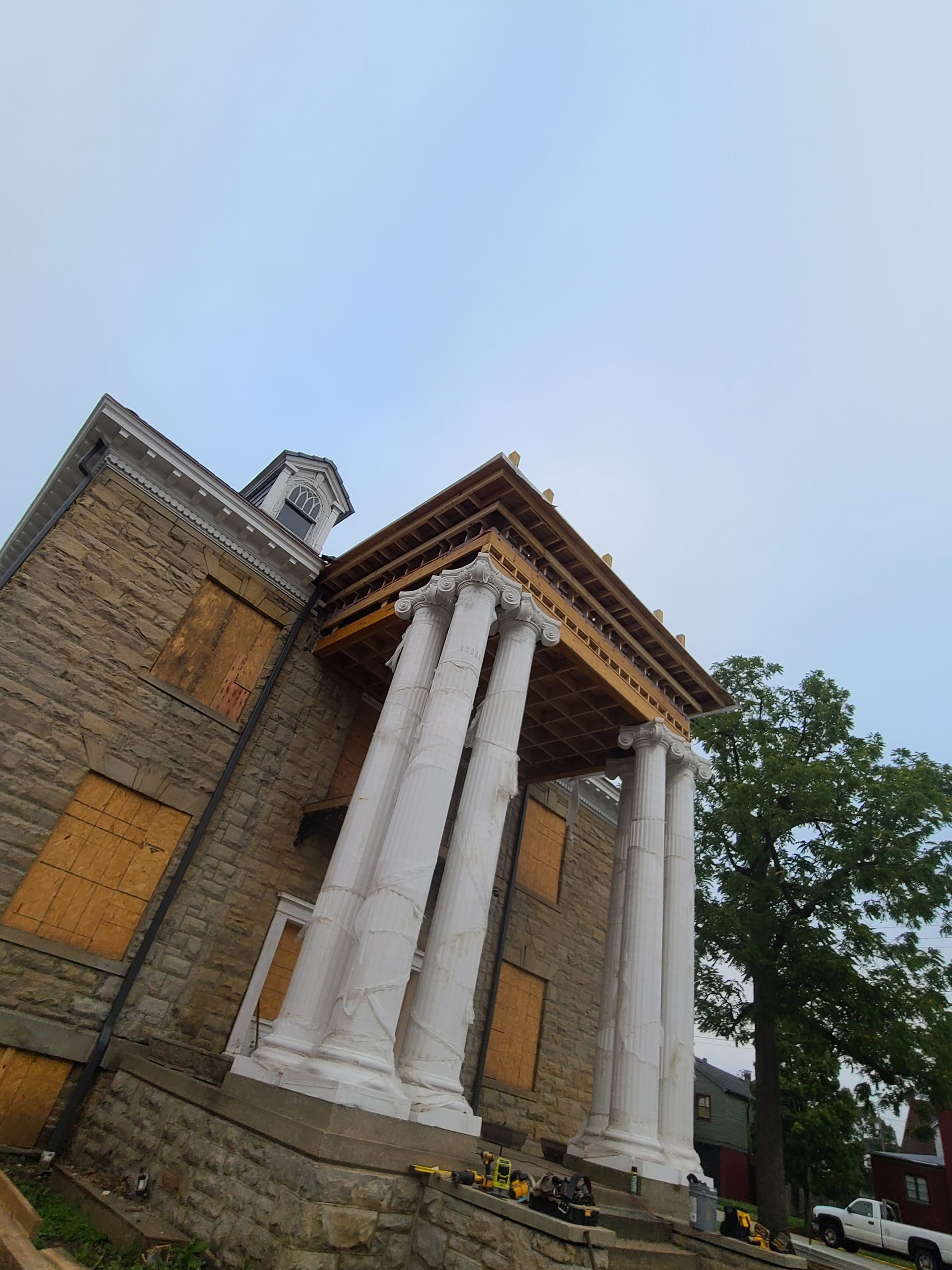 Stone building with large white columns under construction; boarded windows, cloudy sky.