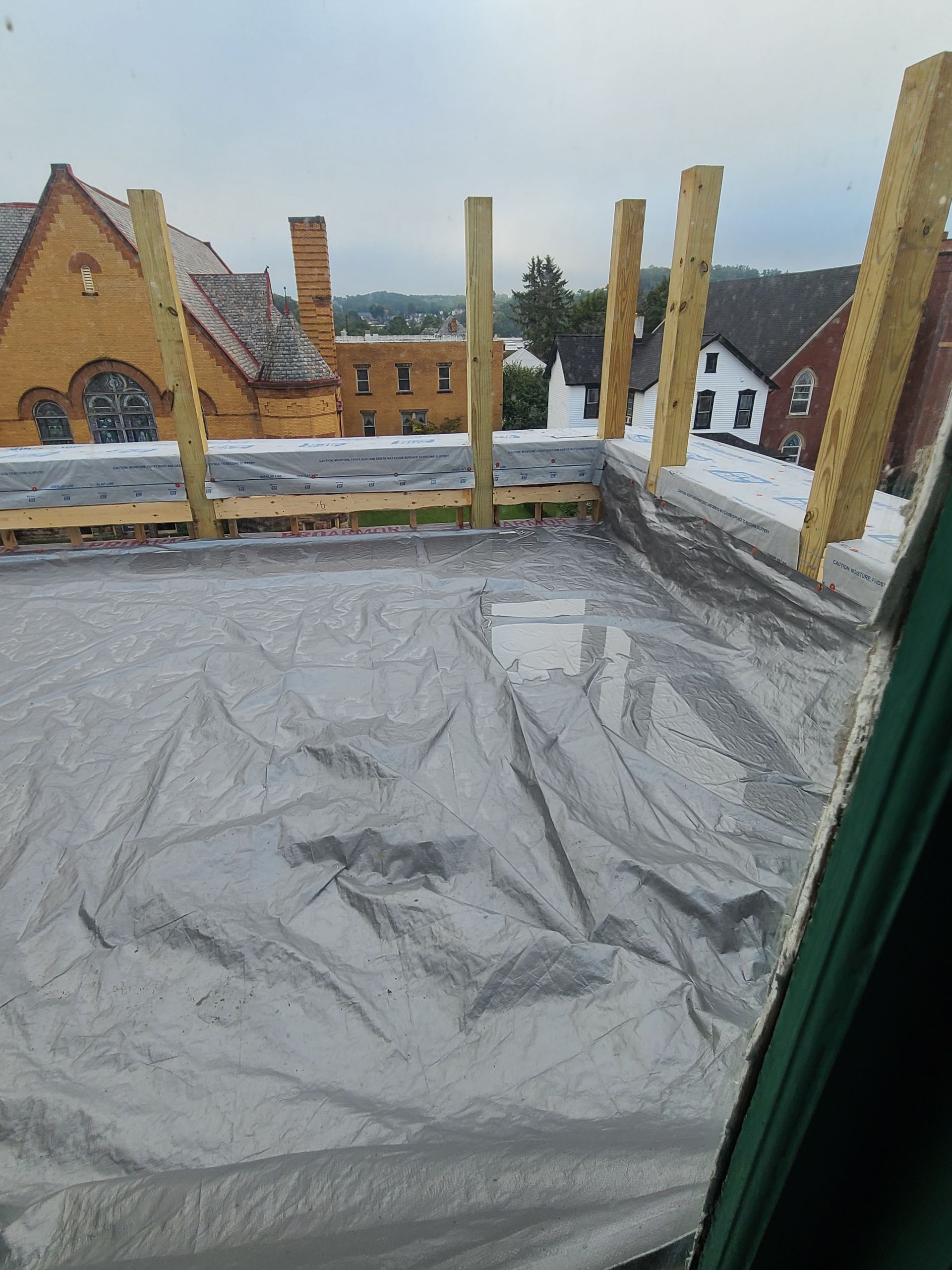 Rooftop construction with wooden beams and a silver tarp. City buildings visible in the background.