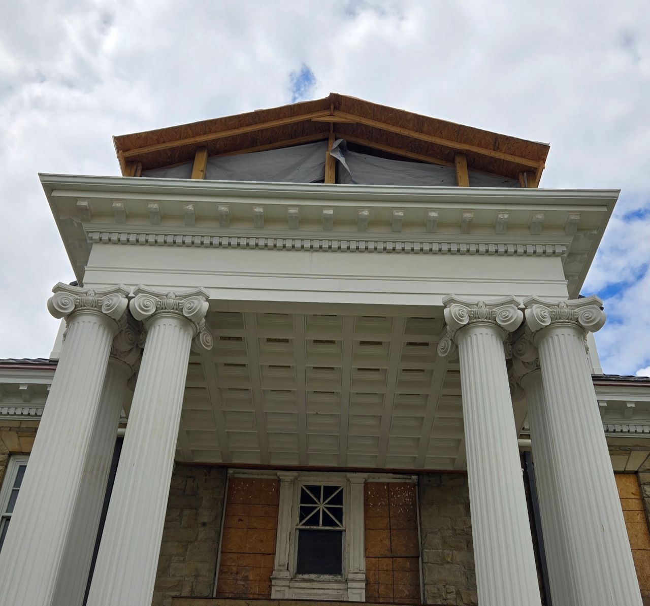 White columns support a roof with a wooden frame against a cloudy sky. The building is beige with a boarded-up window.