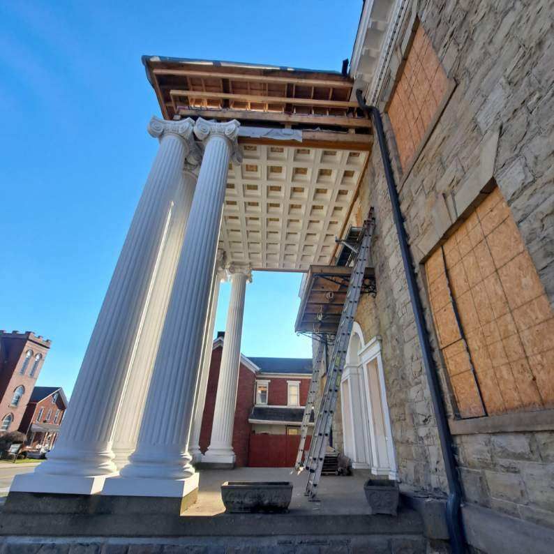 Building facade with white columns; scaffolding and ladder present, with repairs in progress. Boarded-up windows.