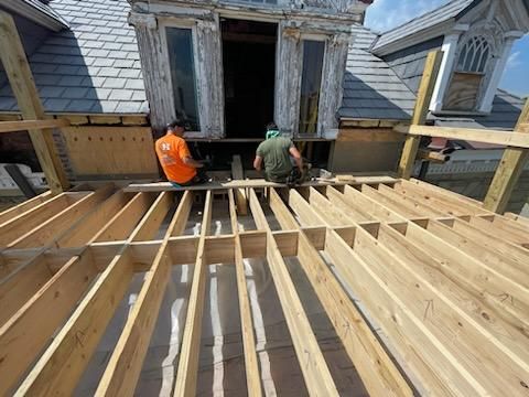 Two workers on a rooftop installing wooden beams. A dormer is visible behind them.