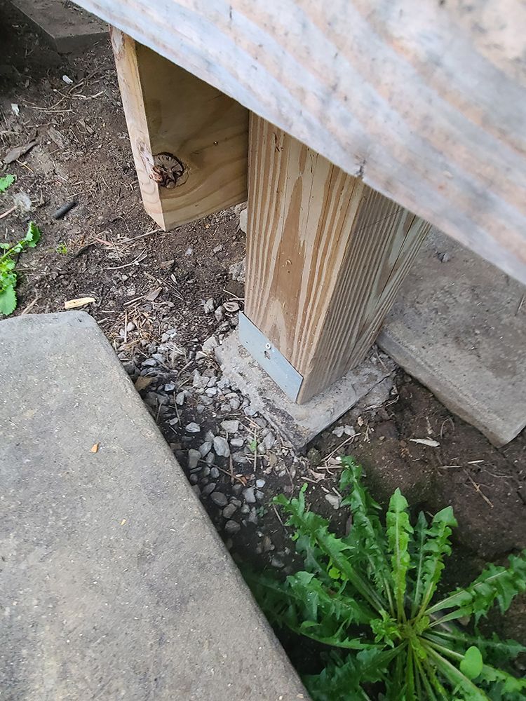 Wooden deck support post on concrete base with gravel and weeds nearby.