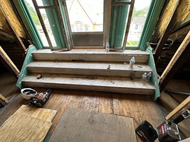 Interior view of attic stairs leading to a window. The stairs are unfinished and dirty, with tools and debris.