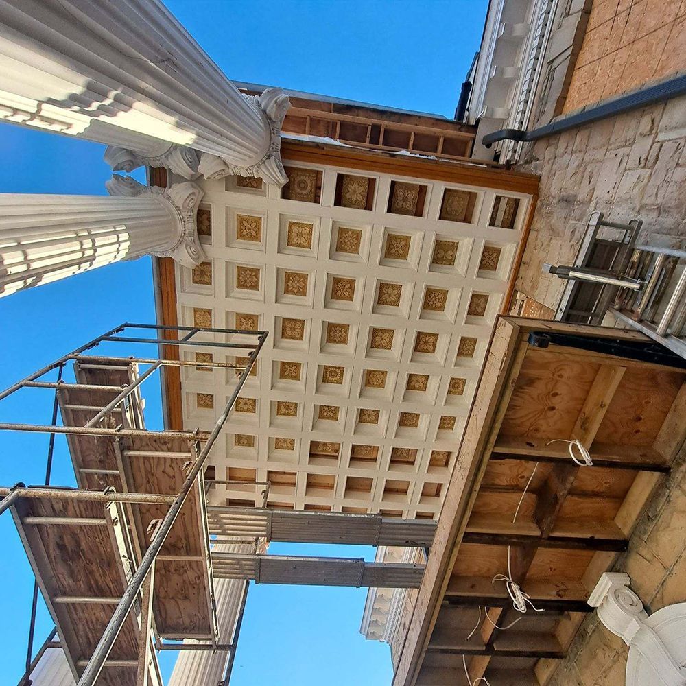 Scaffolding under a decorative ceiling with ornate squares and columns, blue sky.