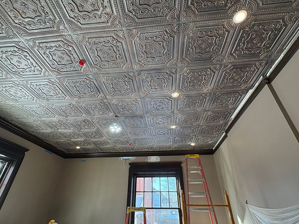 Silver decorative tin ceiling with dark trim and recessed lighting in a room with a window and ladder.