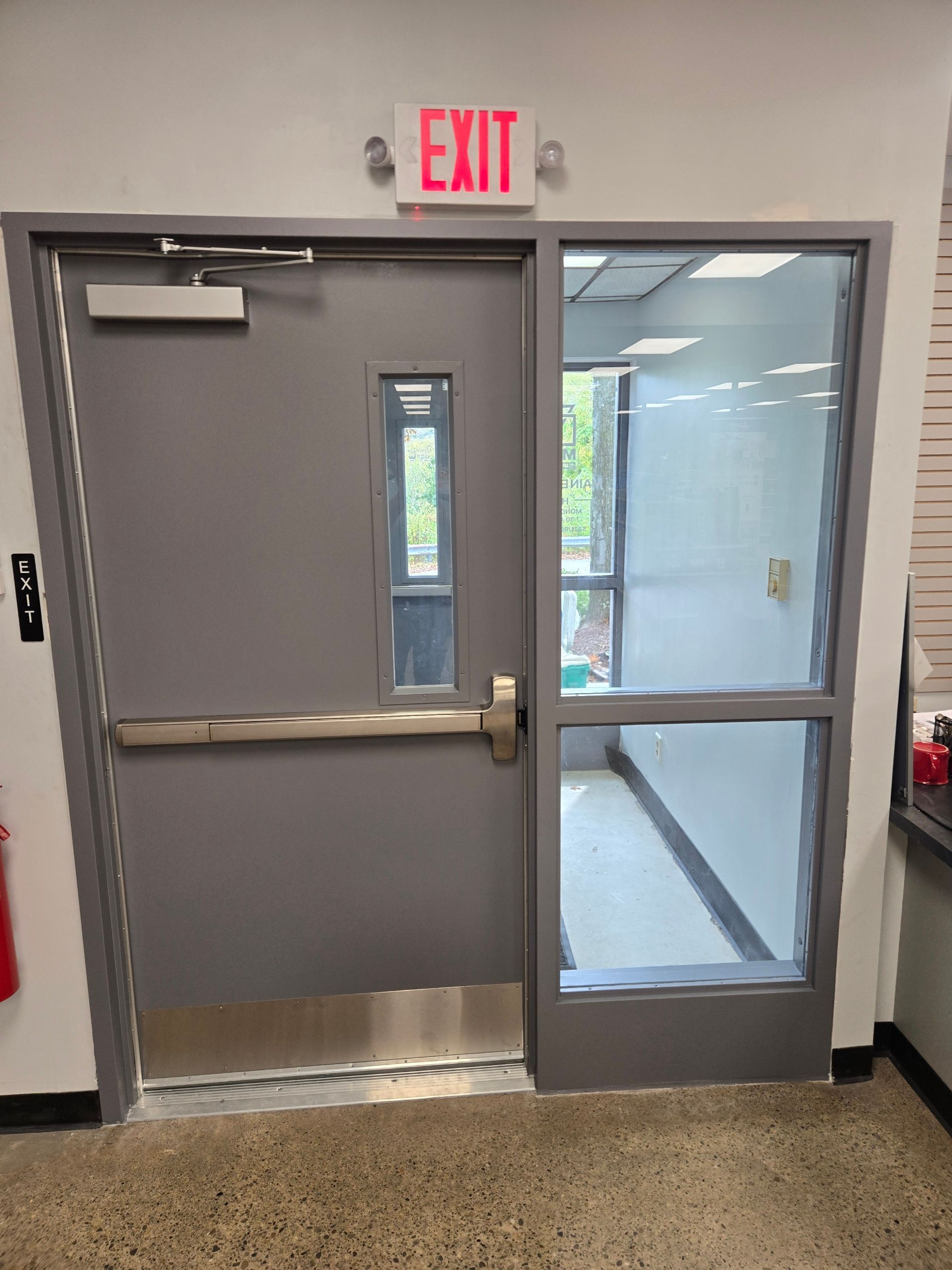 Gray exit door with adjacent glass panel and overhead illuminated 