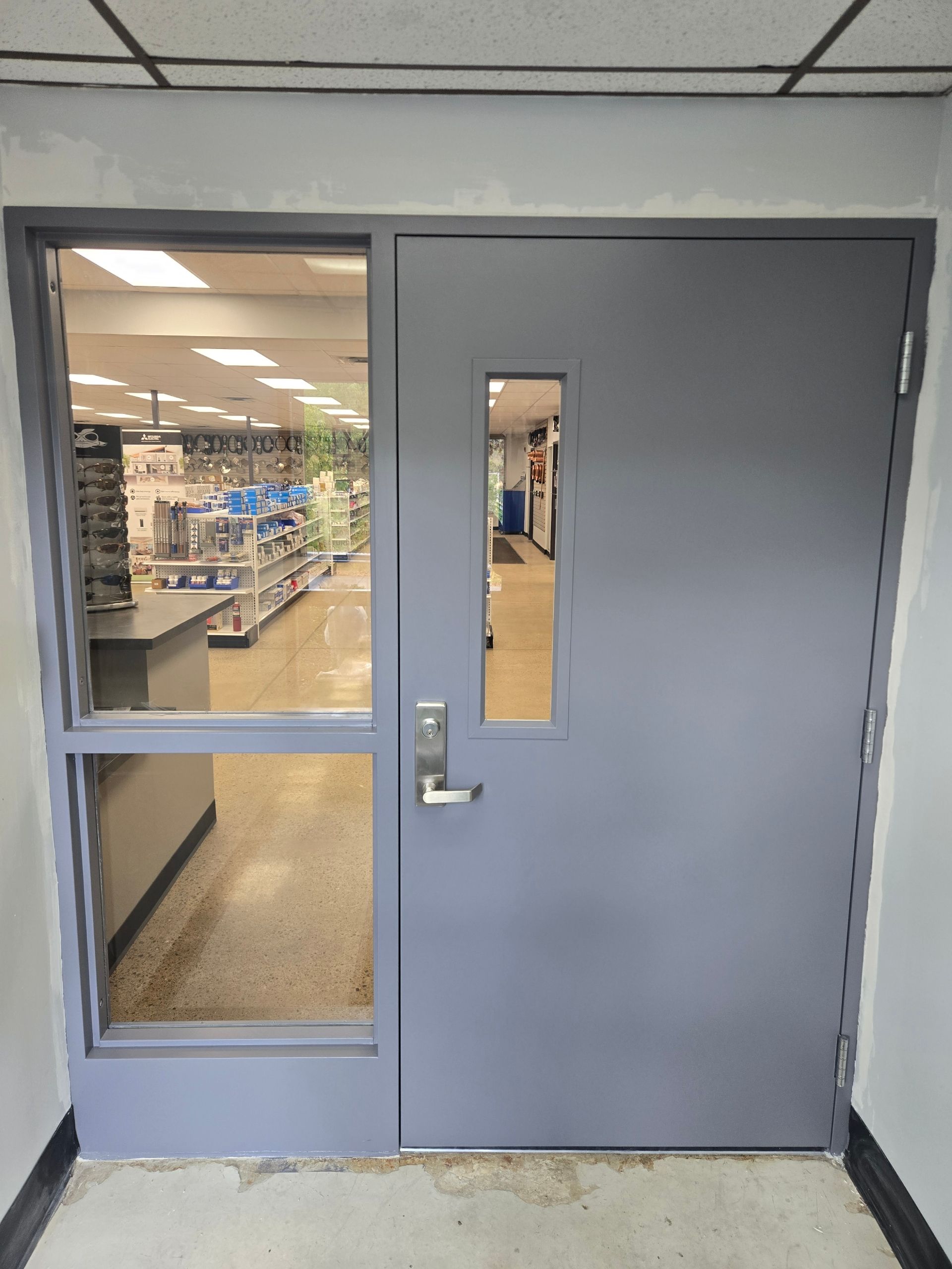 Gray door with sidelight and small window. Inside, a store is visible.