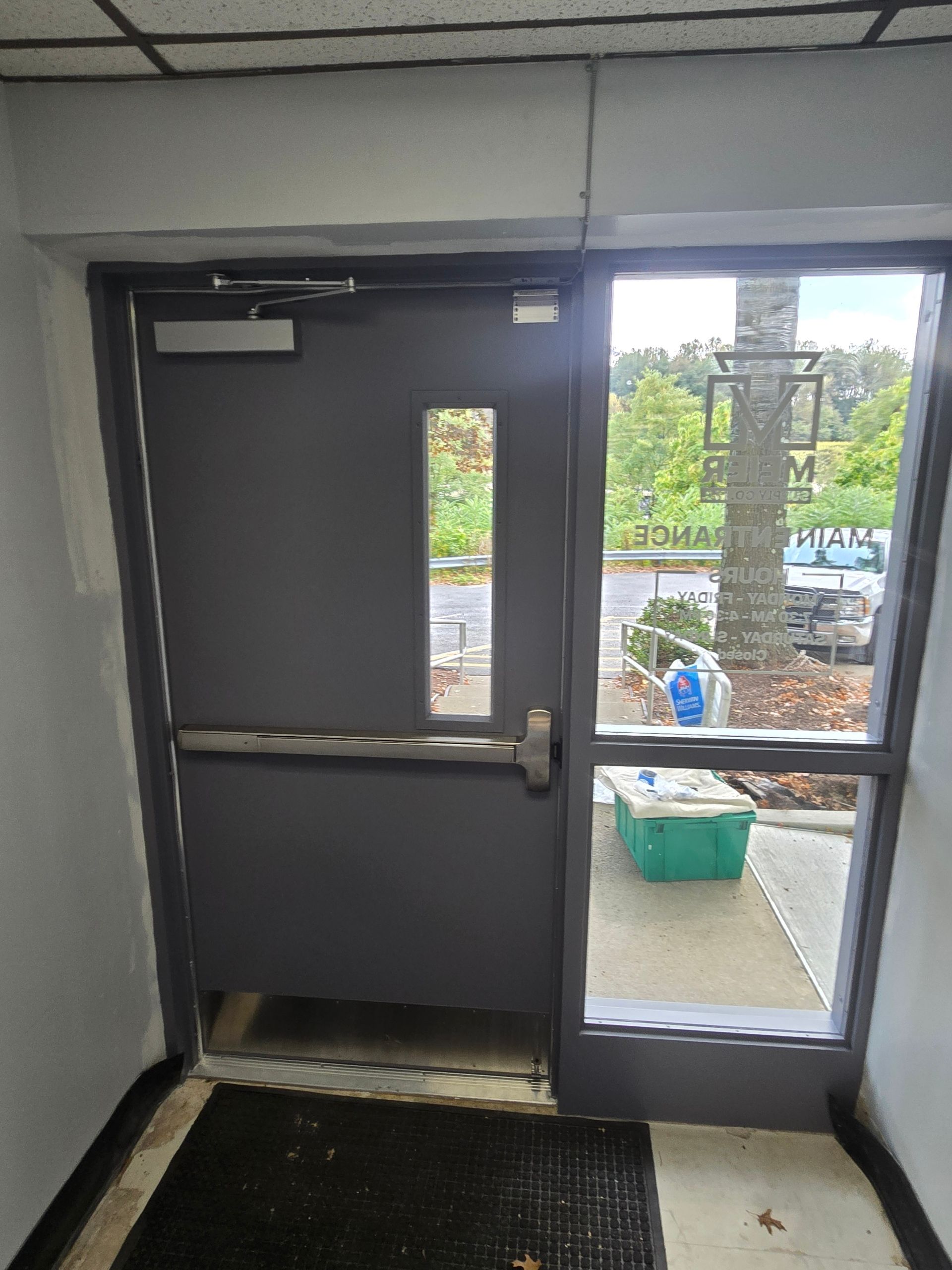 Gray door with window, exit bar, and sidelight. Leads outside, near trash can and tree.