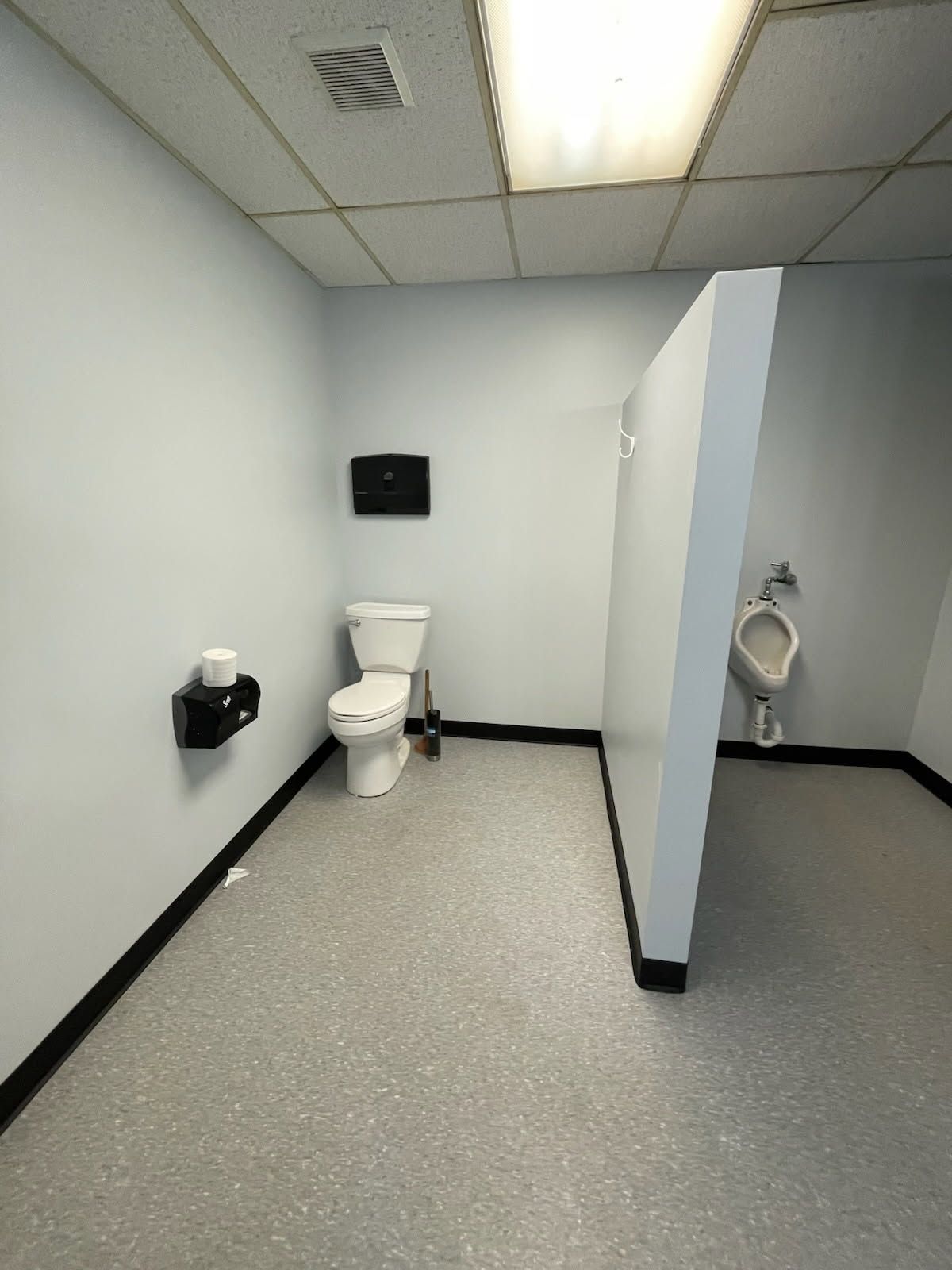 Public restroom with gray walls, toilet, and urinal. Gray and black floor.