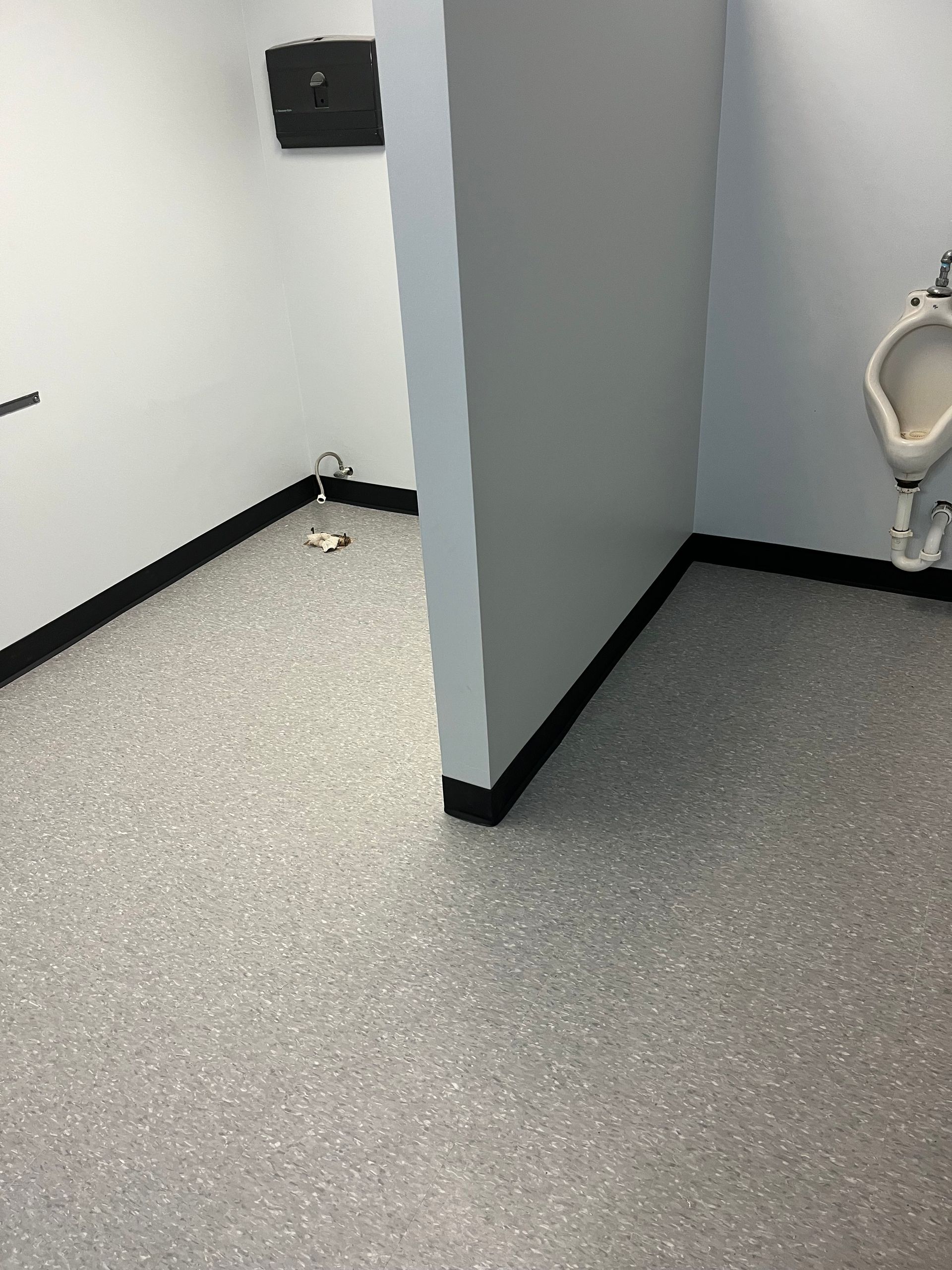A public restroom with gray speckled flooring, white walls, a urinal, and a partition.