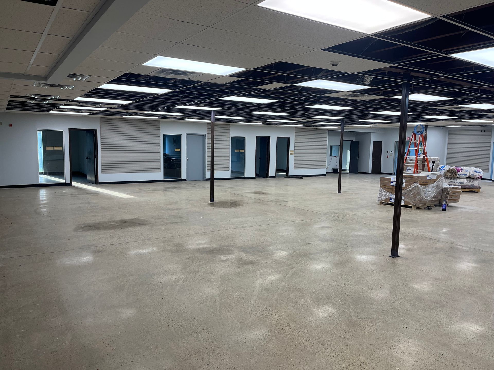 Empty commercial interior, concrete floor, several open doorways, black poles, and ceiling panels.