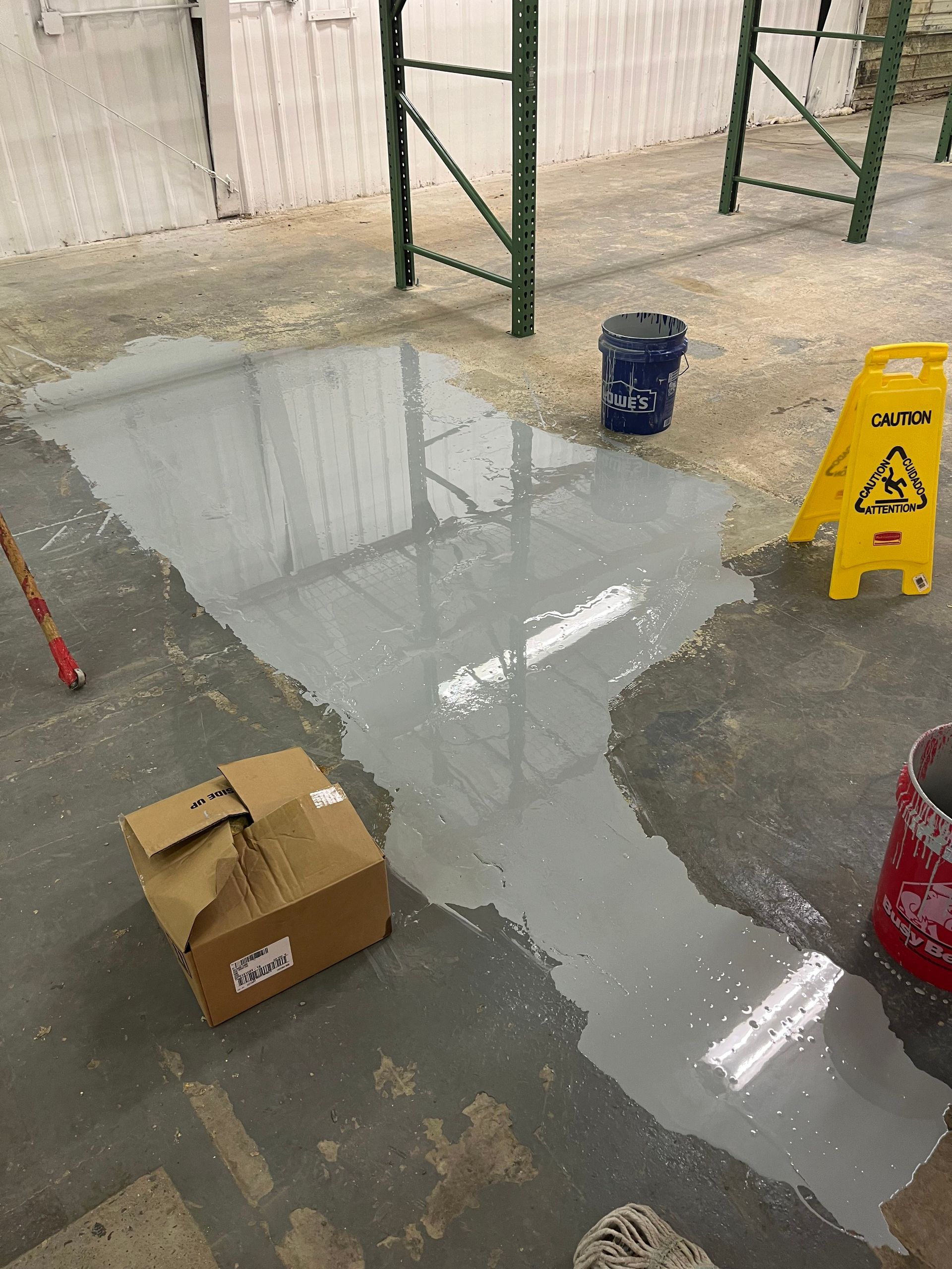 Wet floor with spillage, cardboard box, safety sign, buckets, and shelving in warehouse setting.