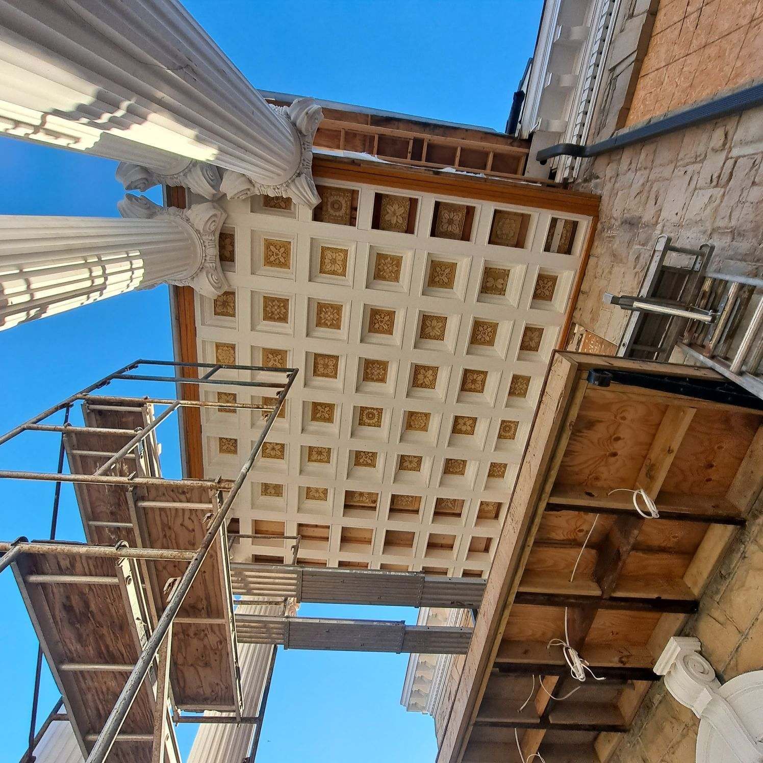 Ornate coffered ceiling being constructed, viewed from below with scaffolding and column.