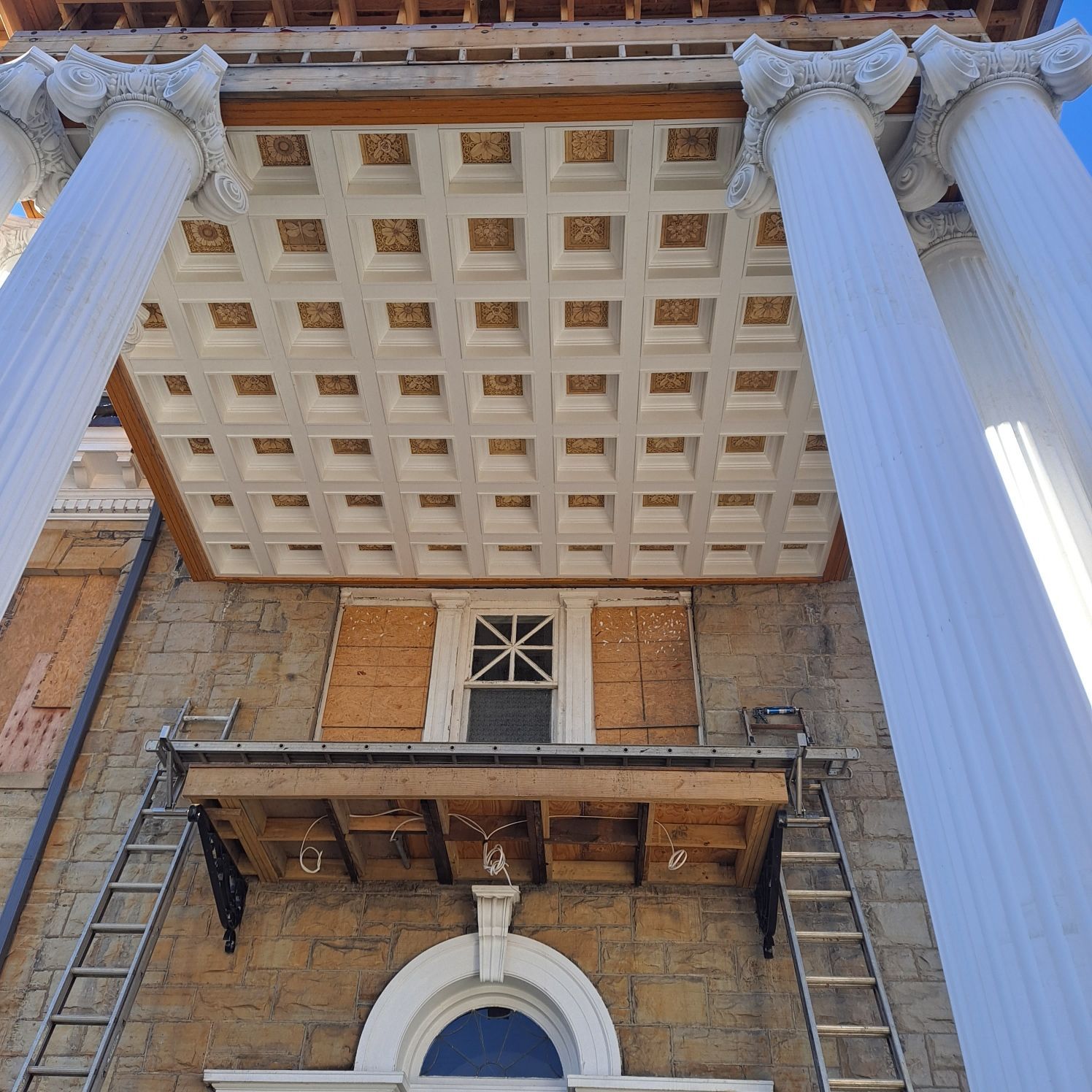 Building facade with large white columns, coffered ceiling, boarded window, and scaffolding.