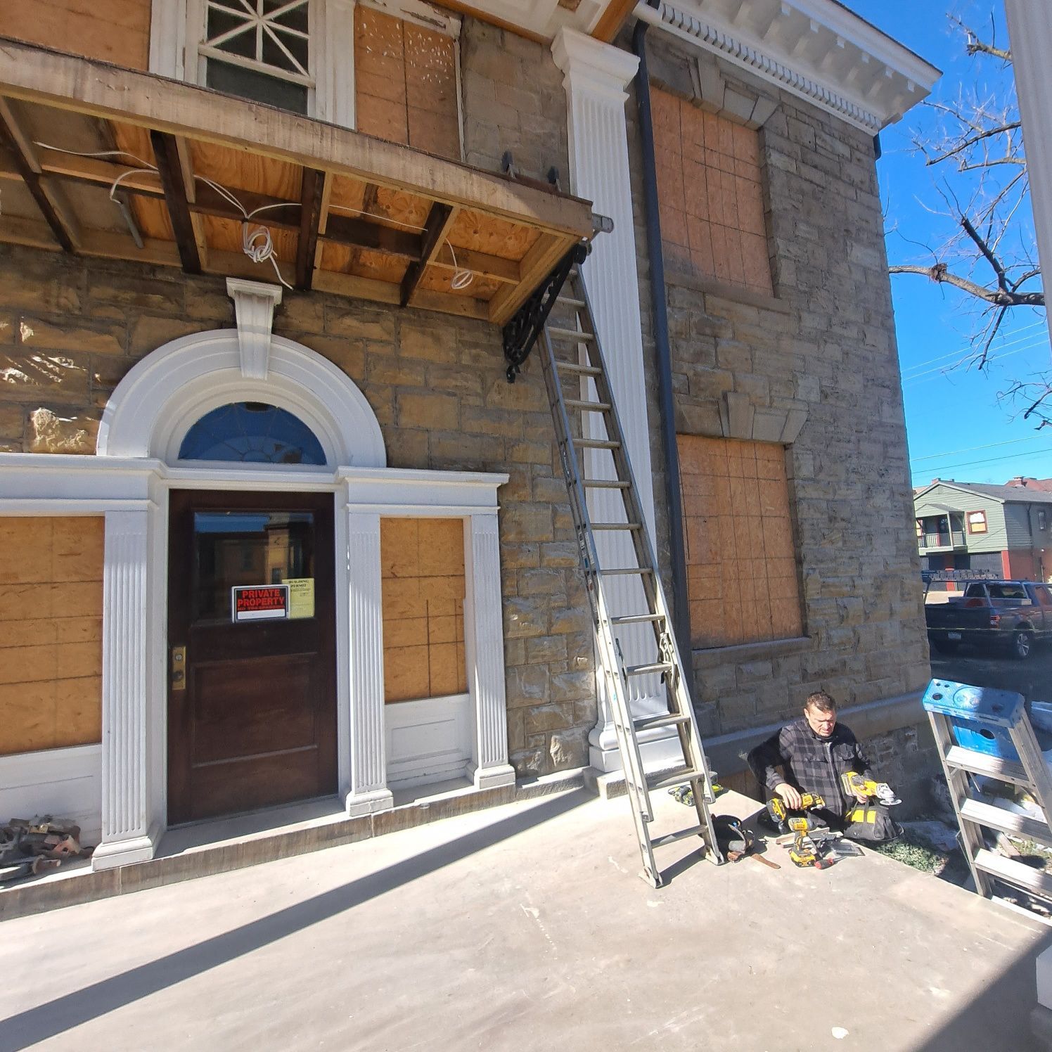 A person working on the facade of a building with boarded windows, ladder propped up.