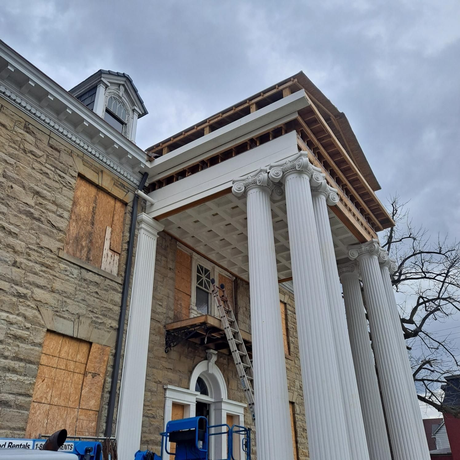 Building facade with white columns and construction work underway; boarded windows.