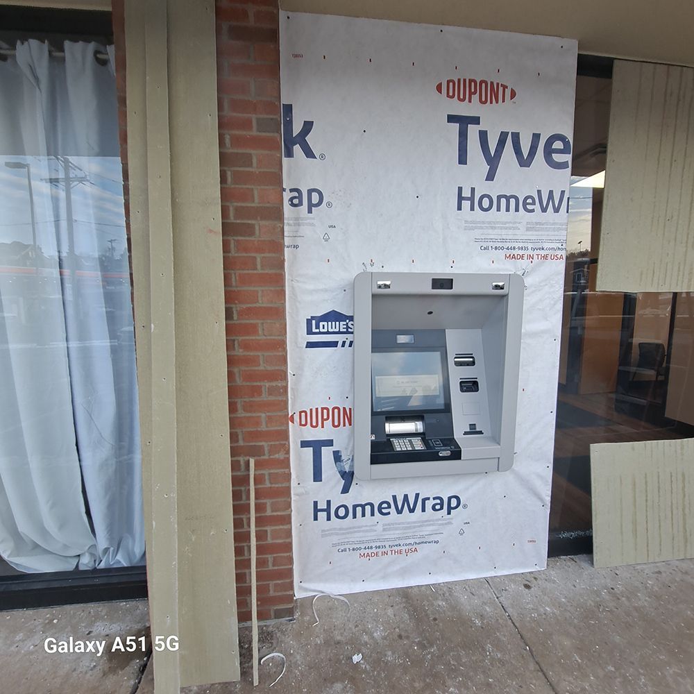 ATM installed on a building with Tyvek wrap and brick, curtains visible through the window.