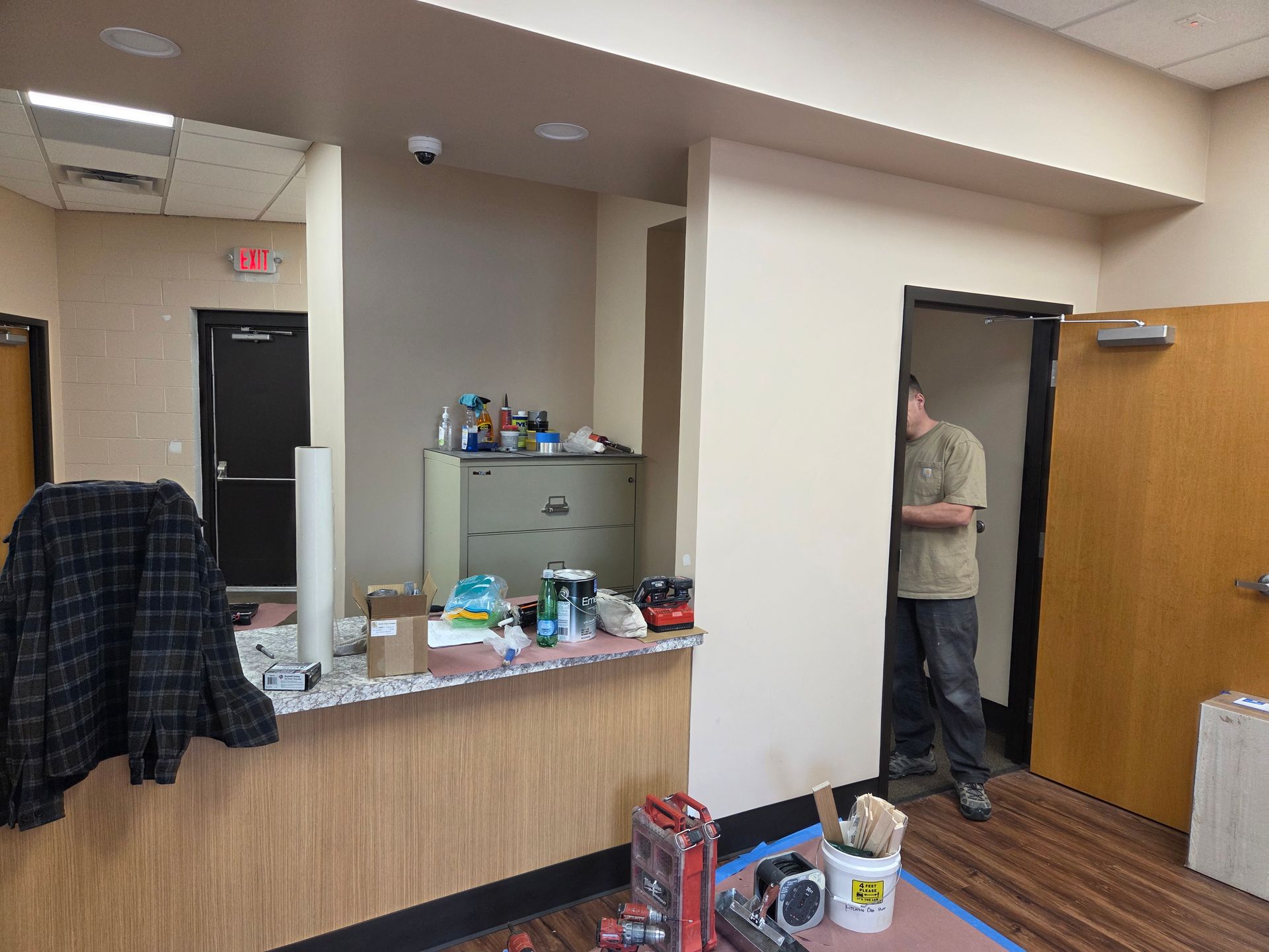 A person peers from a doorway. A counter cluttered with items sits in the foreground. Office interior.