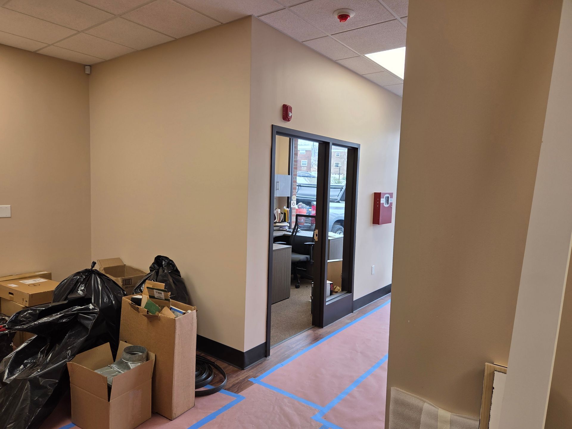 Empty office hallway with boxes, trash bags, and a doorway. Beige walls and red fire alarm visible.