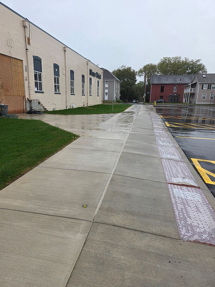 Exterior view of a wet sidewalk next to a building and parking lot, overcast sky.