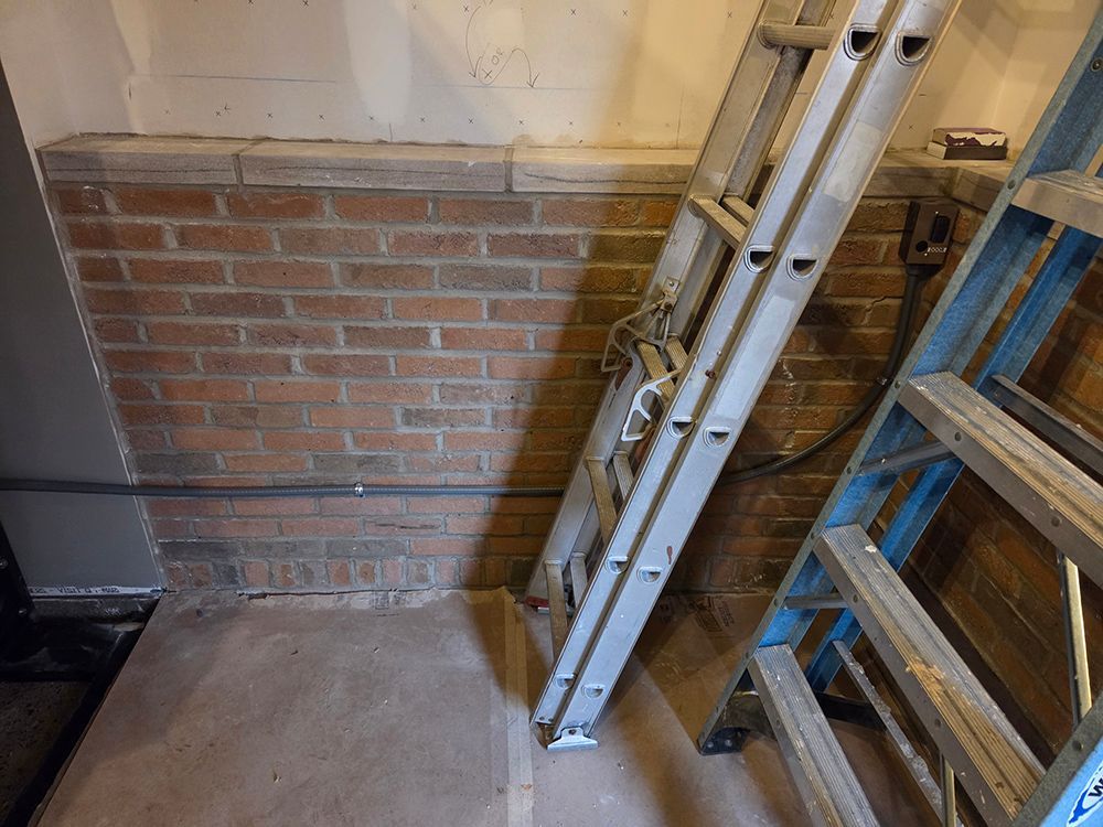 Ladder leaning against a brick wall in a basement, alongside another ladder; gray floor.