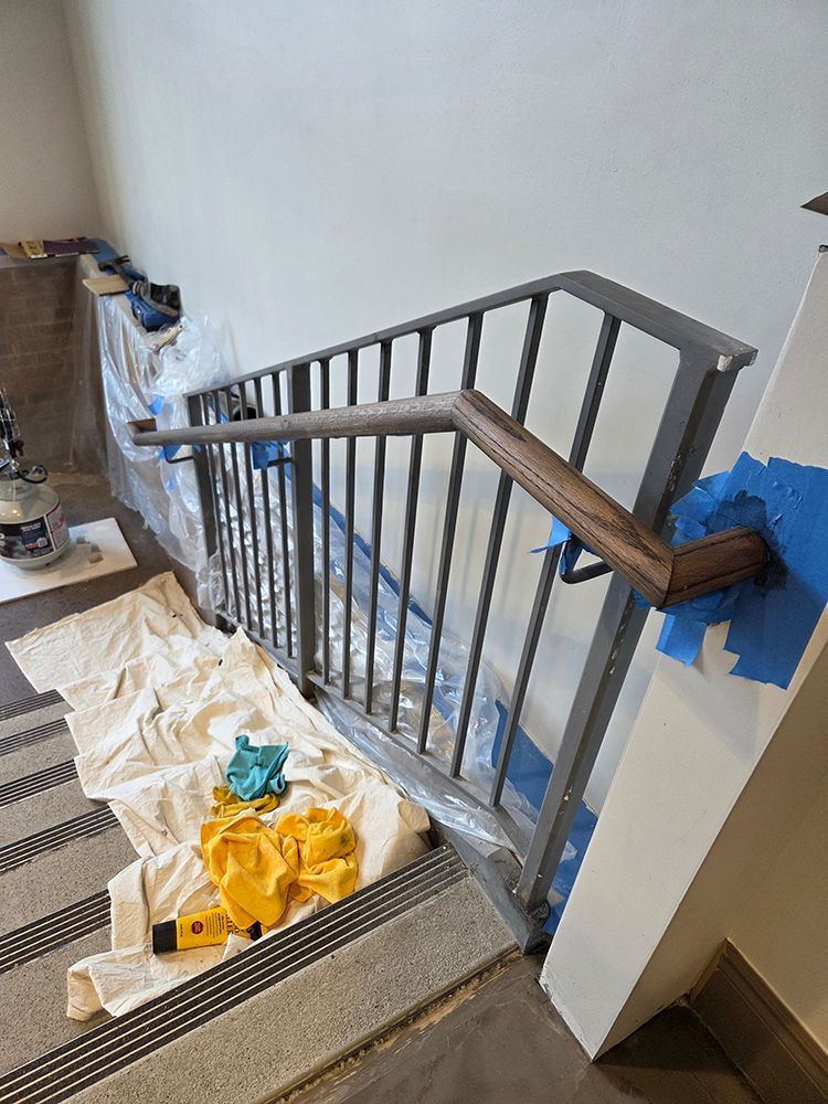 Staircase handrail partially painted, with plastic sheeting covering steps and tools present.