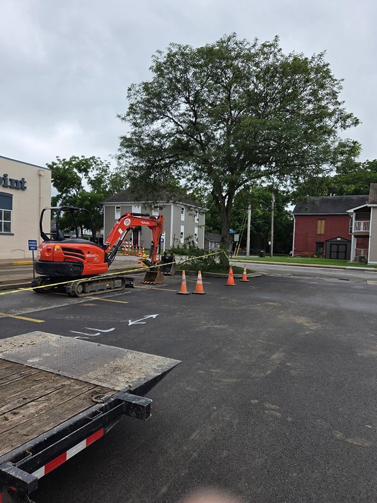 Orange excavator, cones, and caution tape in a parking lot, near a tree and buildings.
